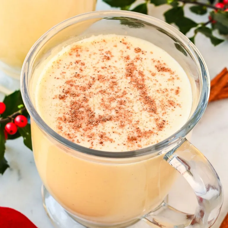 Glass mugs of non-alcoholic eggnog topped with cinnamon and nutmeg, garnished with holly and cinnamon sticks for Christmas.