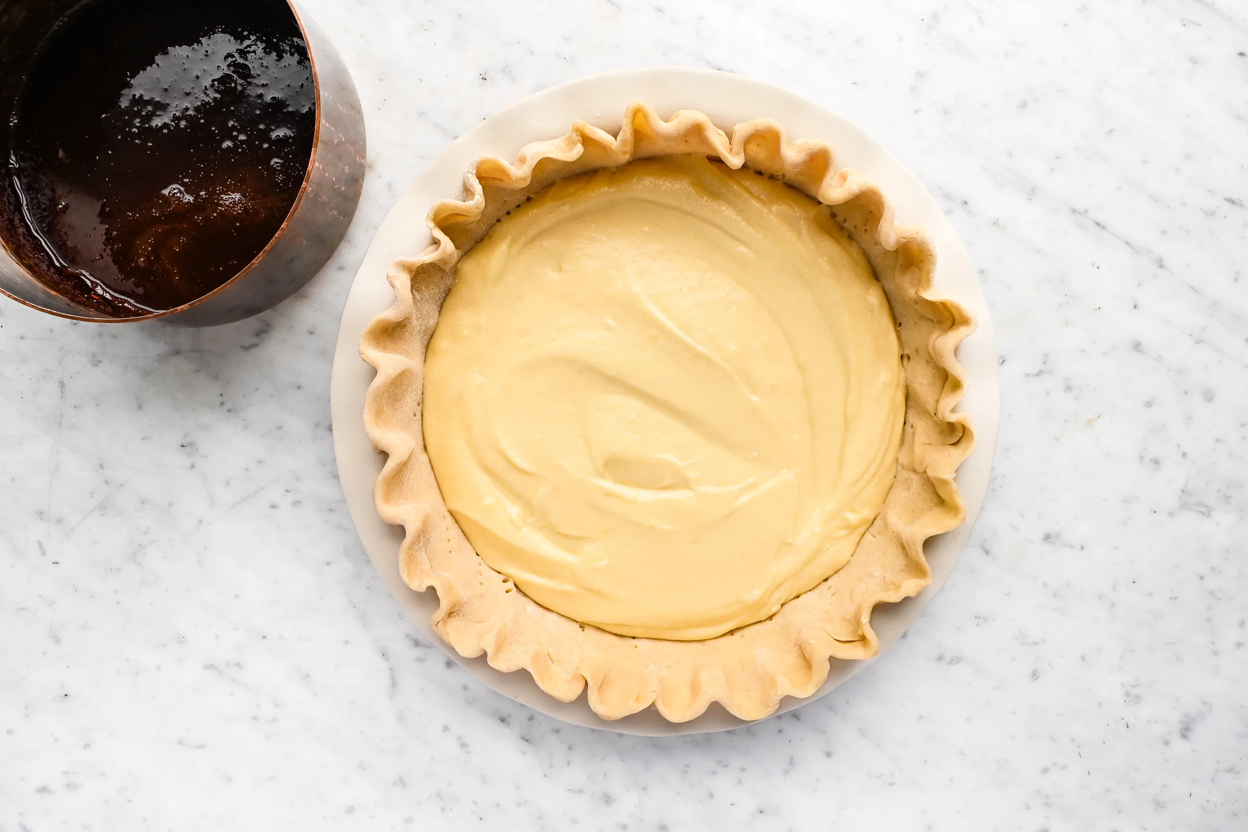 Pie crust filled with smooth vanilla batter and a saucepan of warm chocolate syrup beside it, ready to pour for making classic Funny Cake.