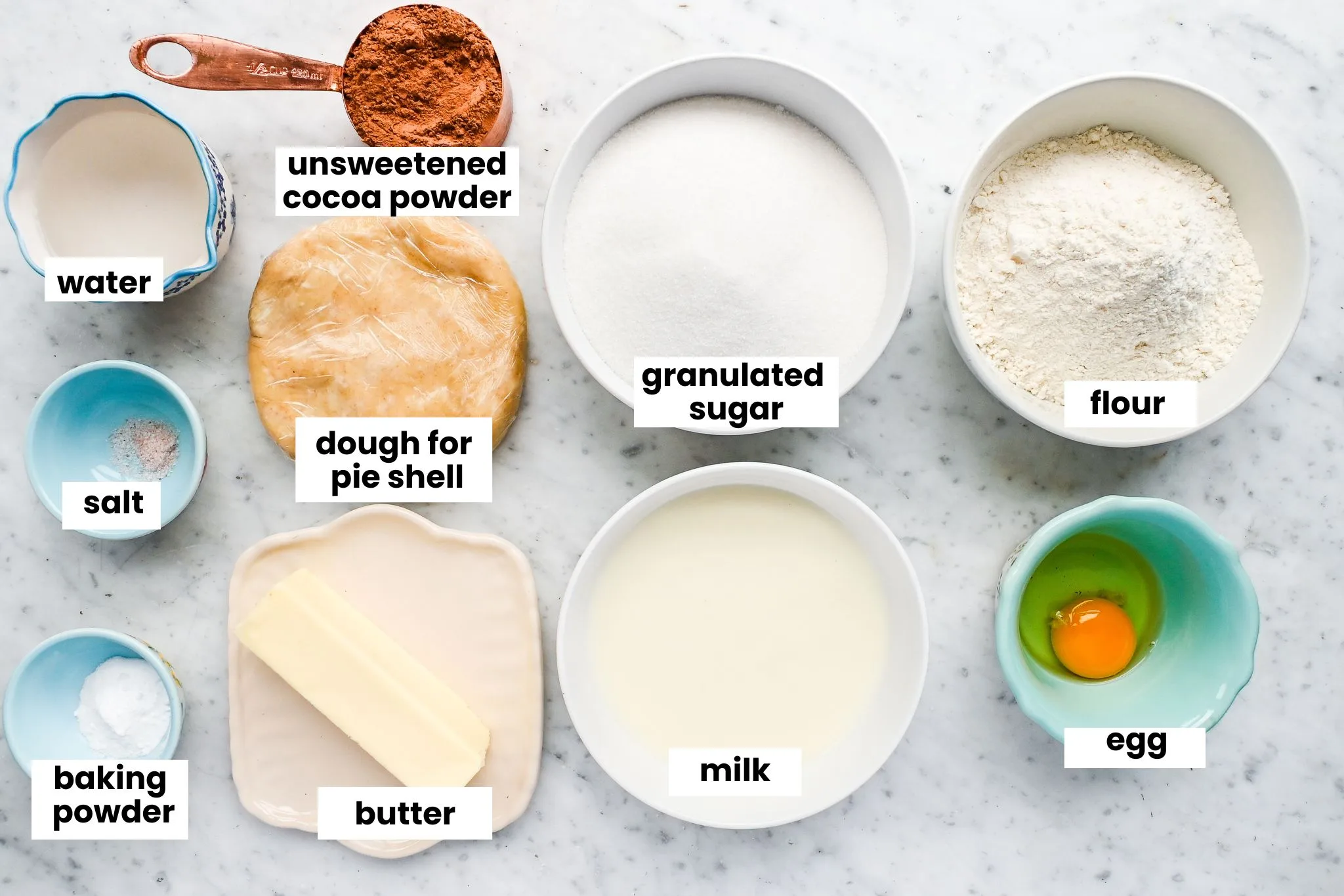 Ingredients for Pennsylvania Dutch Funny Cake including cocoa, flour, sugar, butter, egg, and milk arranged on a marble countertop.