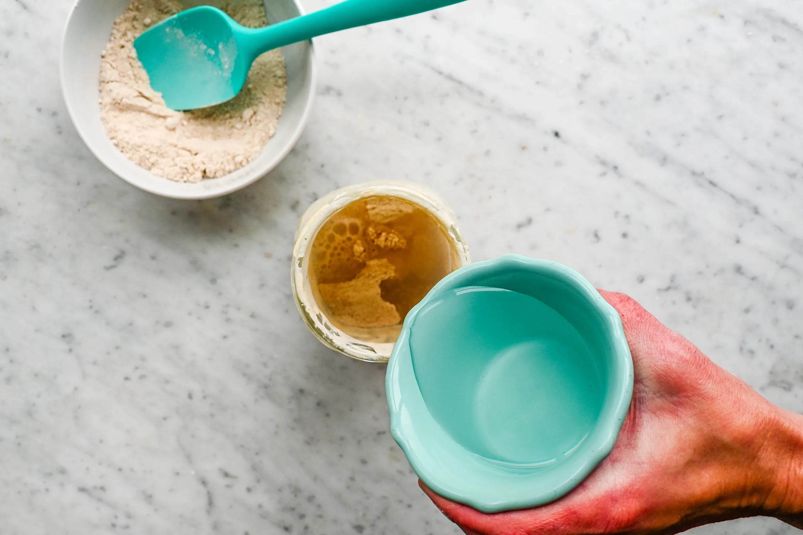 Hand holding a small turquoise bowl of water next to a jar of active sourdough starter and a bowl of freshly milled flour.