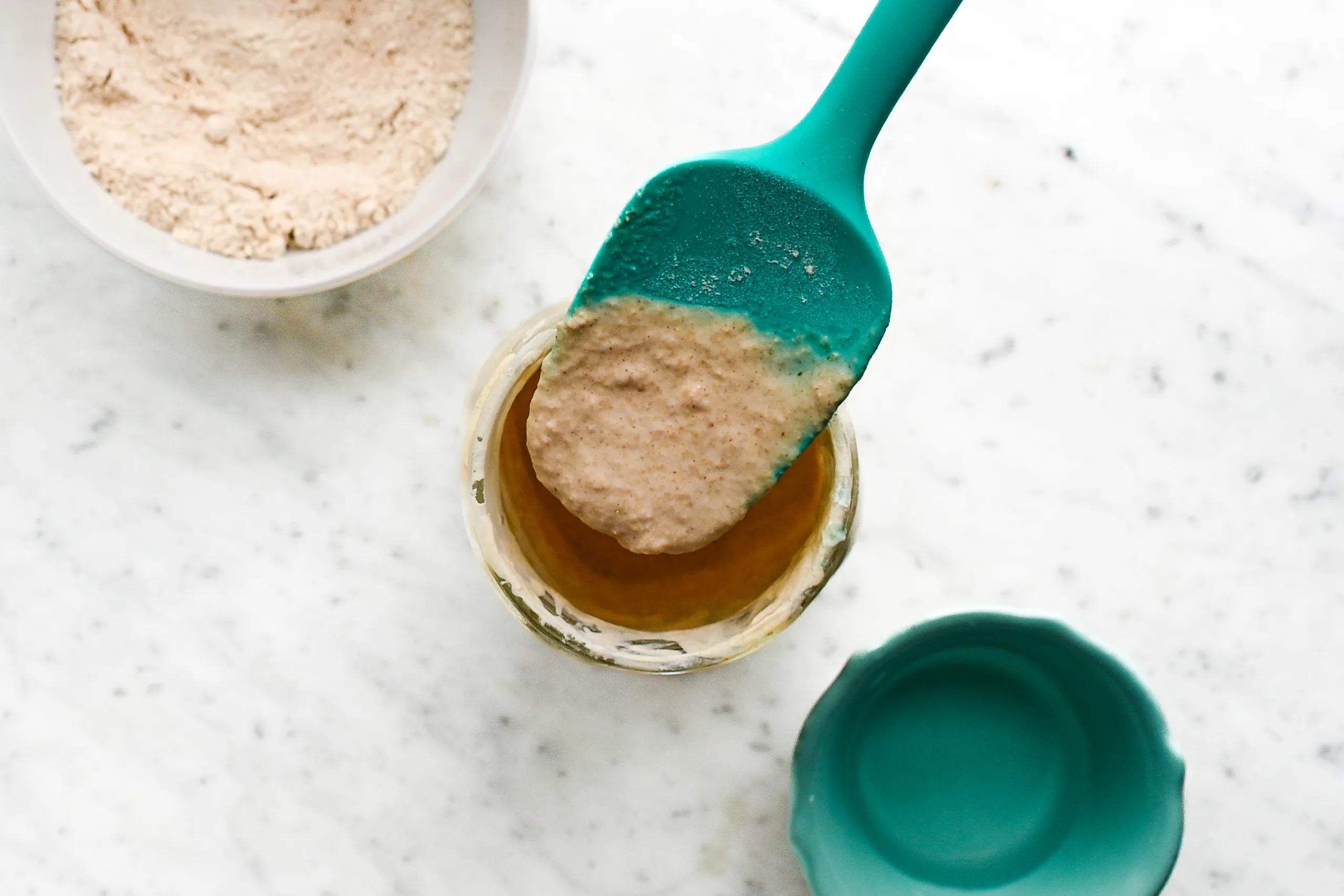 Spatula lifting freshly fed sourdough starter made with whole wheat or milled flour from a glass jar.