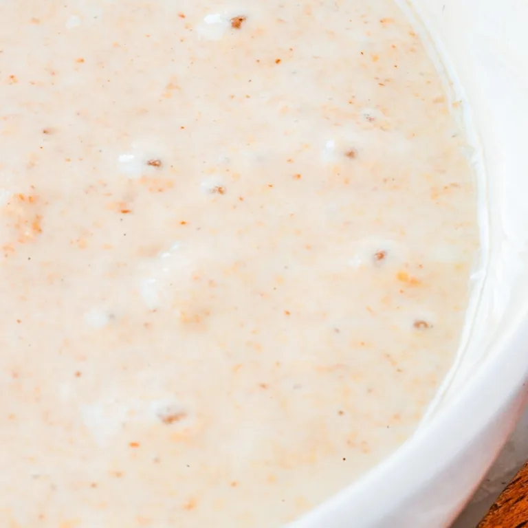 Creamy, lightly speckled sourdough starter in a white bowl, showing small bubbles forming across the surface as it ferments.
