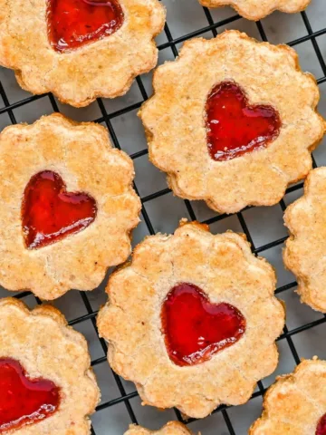 “Golden jam-filled pie crust cookies cooling on a wire rack, each one shaped with scalloped edges and a bright red heart of jam in the center.