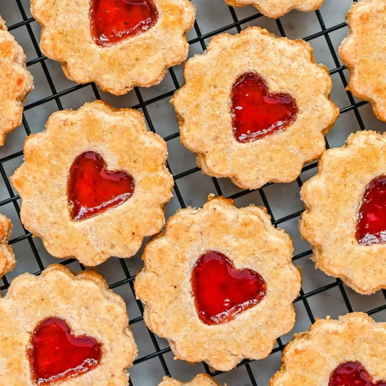 “Golden jam-filled pie crust cookies cooling on a wire rack, each one shaped with scalloped edges and a bright red heart of jam in the center.
