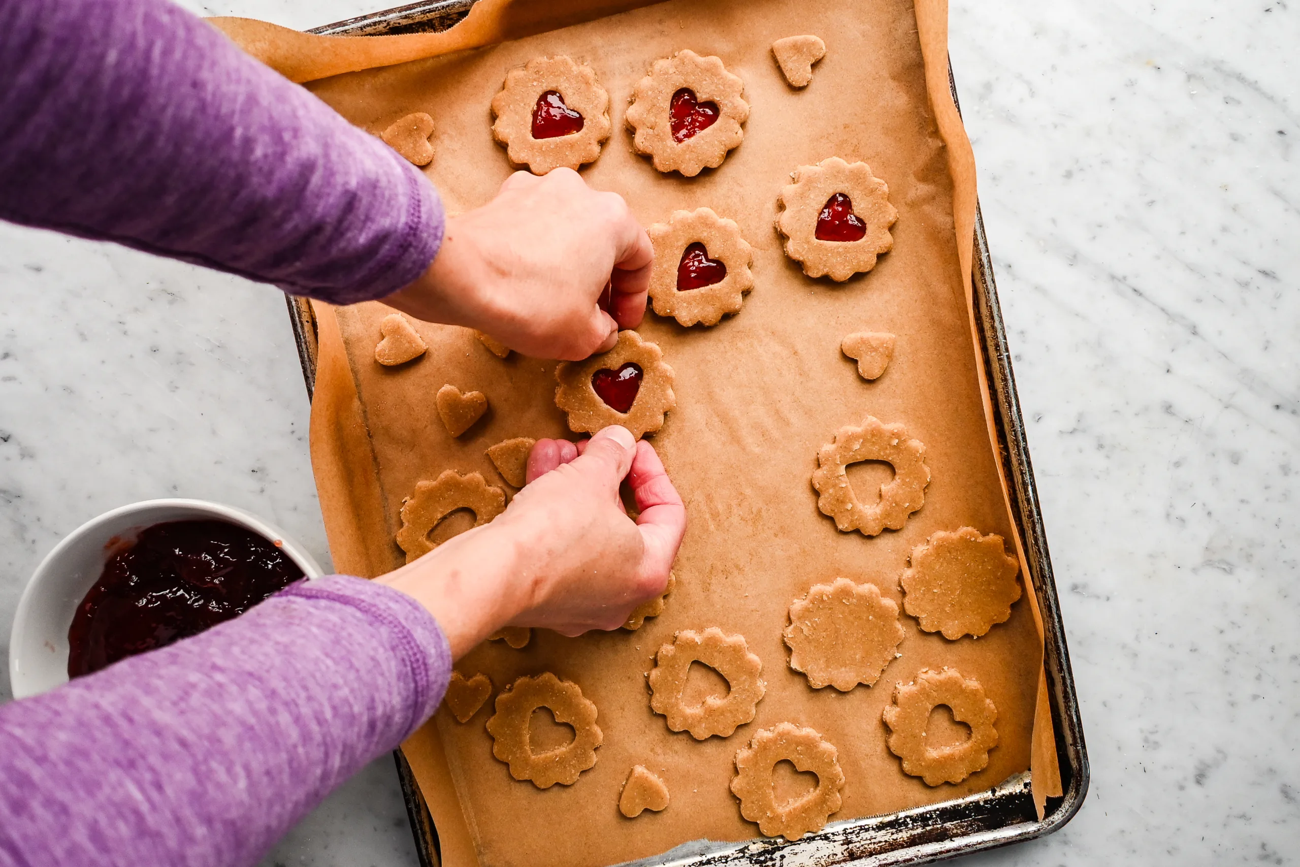 Hands placing cutout cookie tops over small spoonfuls of red jam to assemble jam-filled pie crust cookies on a parchment-lined baking sheet.