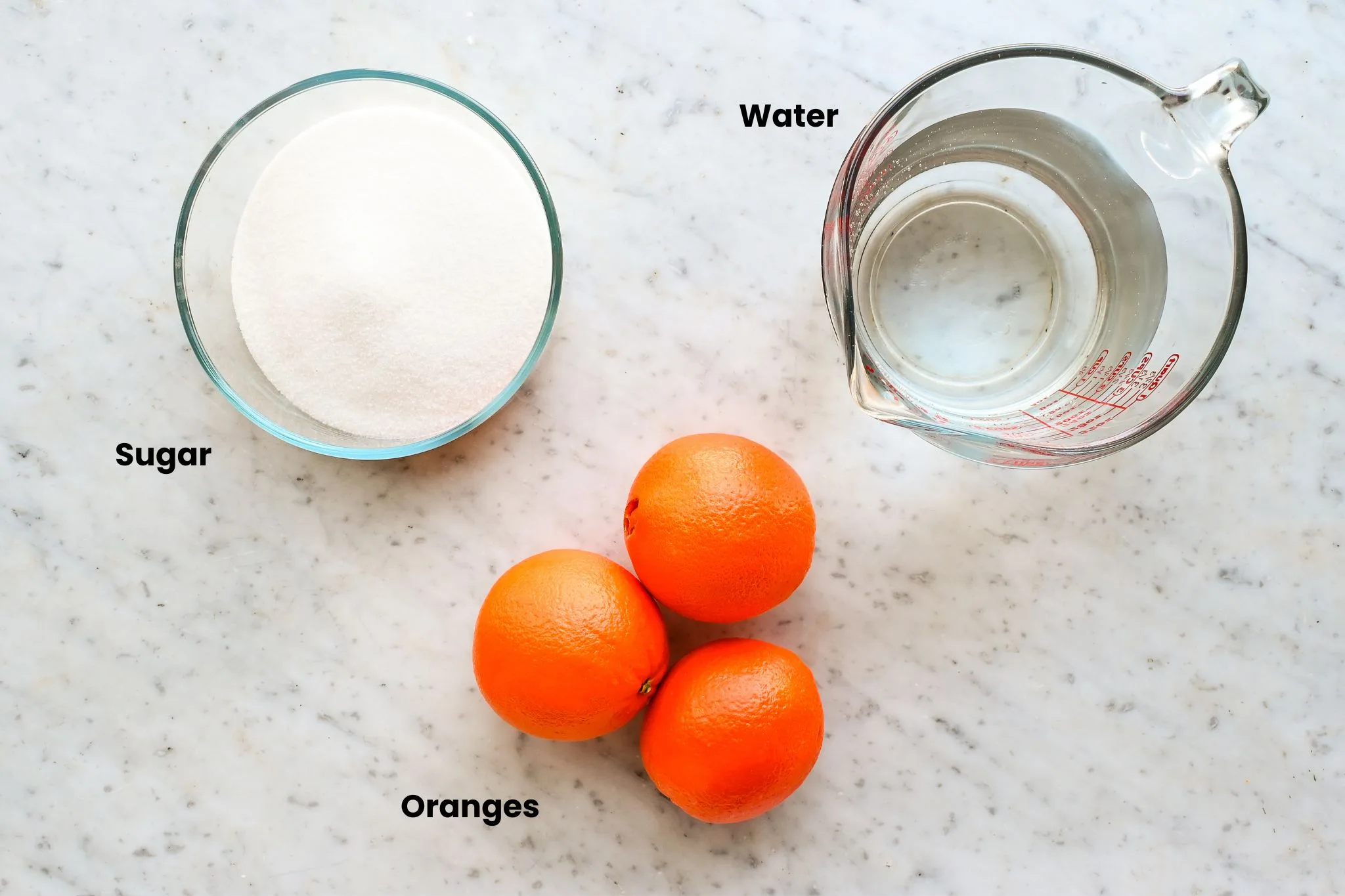 Granulated sugar, water, and whole oranges arranged on a light surface, ingredients measured and ready for making candied orange peels from scratch.
