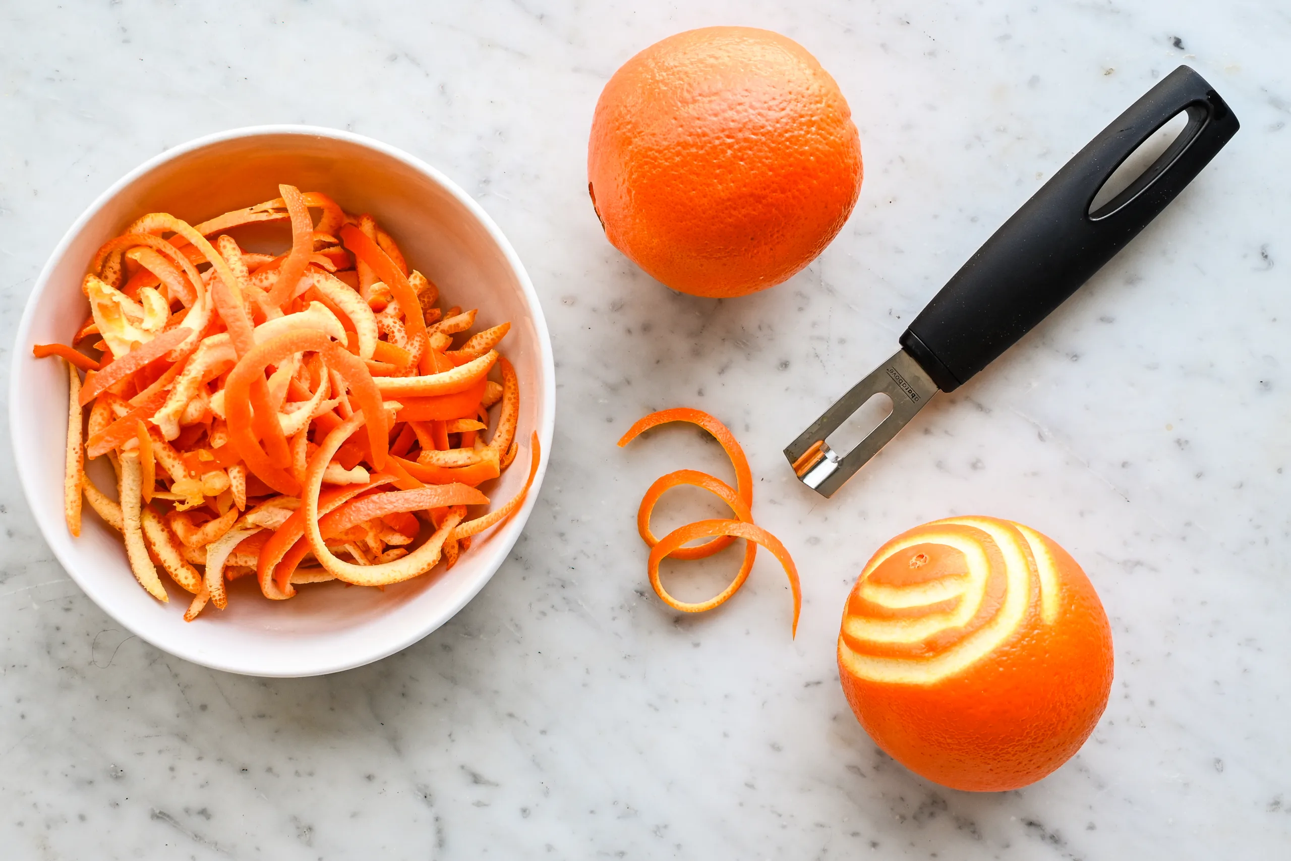 Fresh orange peels curled into thin strips beside a peeler and whole oranges on a marble countertop, showing preparation for homemade candied orange peel.
