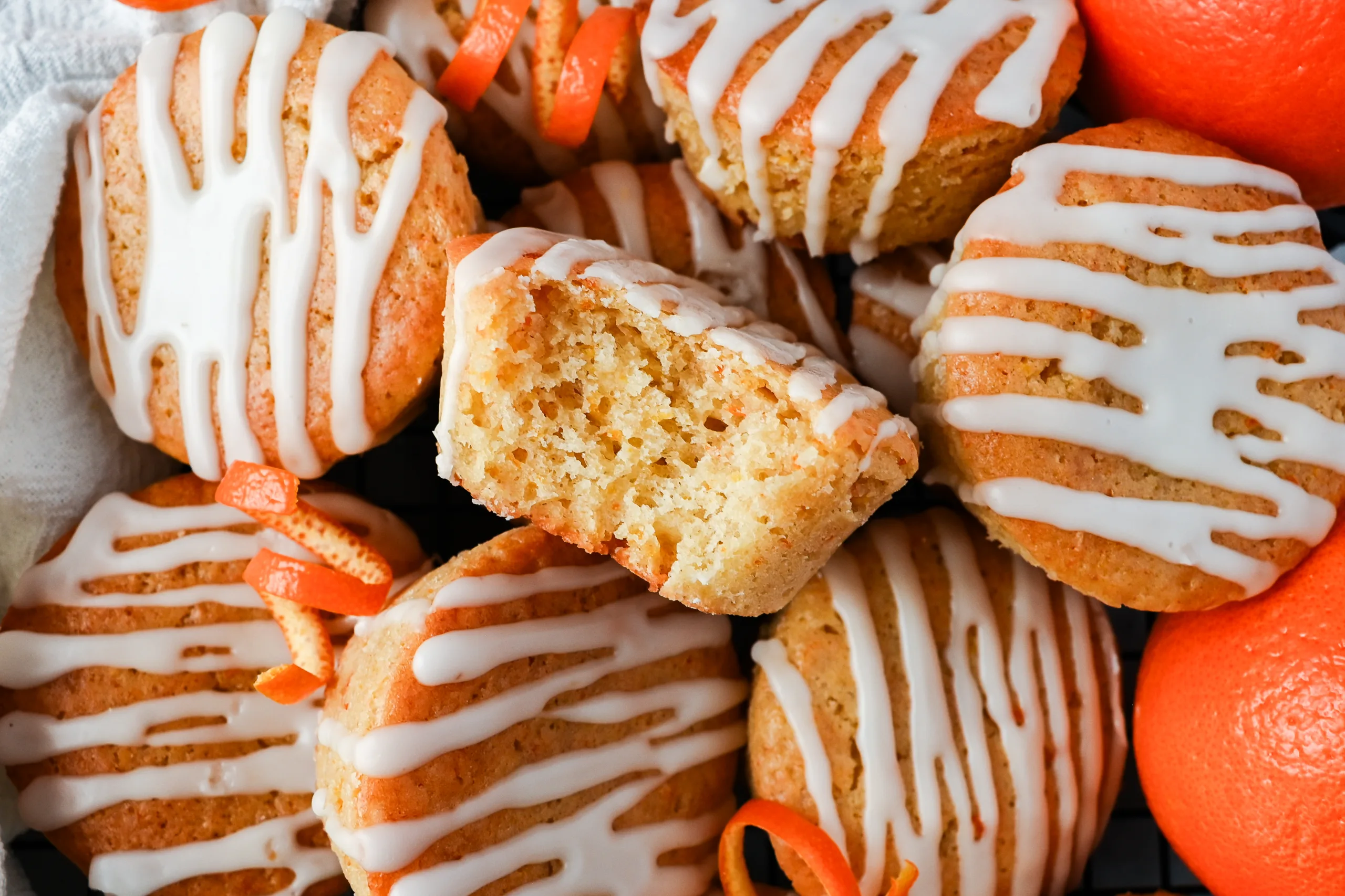 Orange muffins with a white citrus glaze cooling on a wire rack, with one muffin cut open to show the soft crumb inside.
