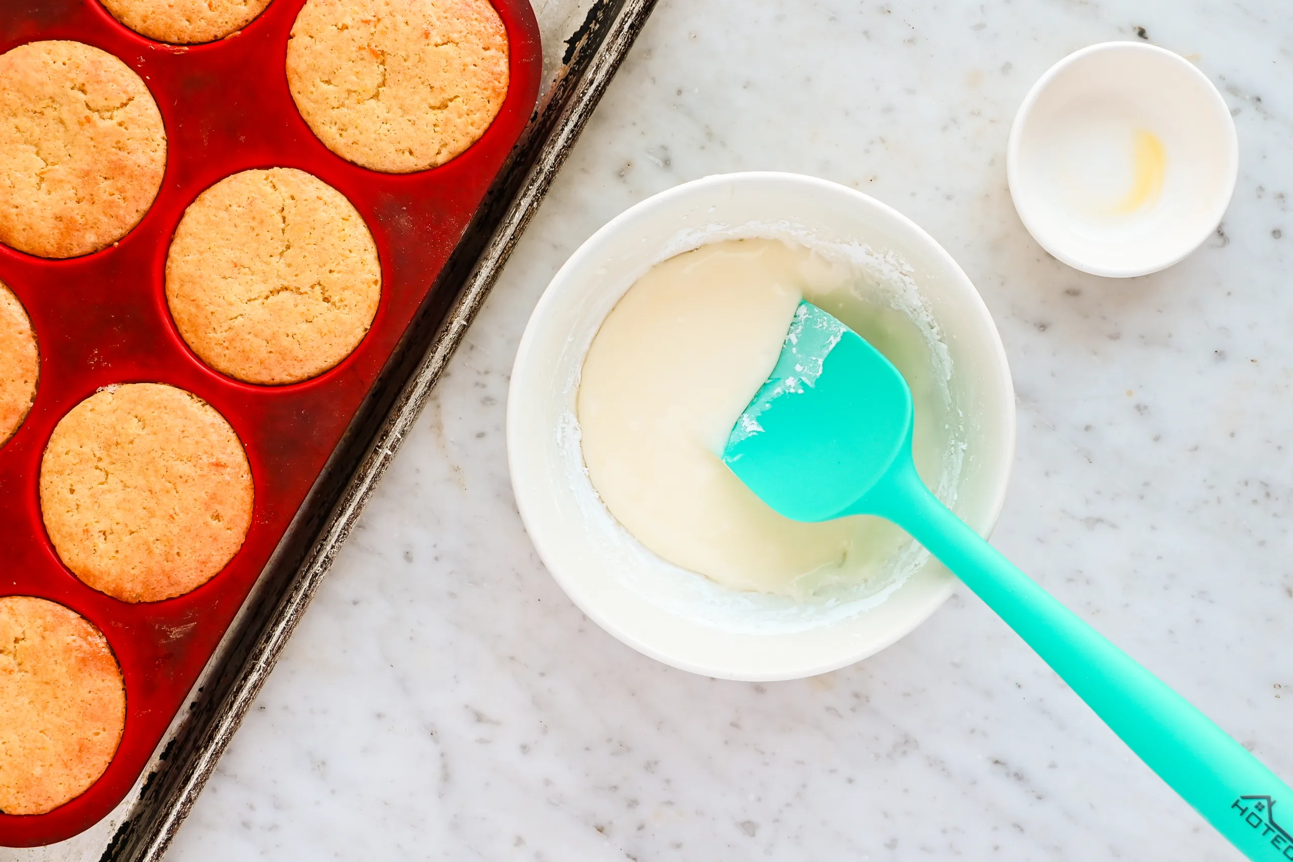 Freshly baked orange muffins in a silicone pan beside a bowl of orange glaze, ready to be spooned over the warm muffins.