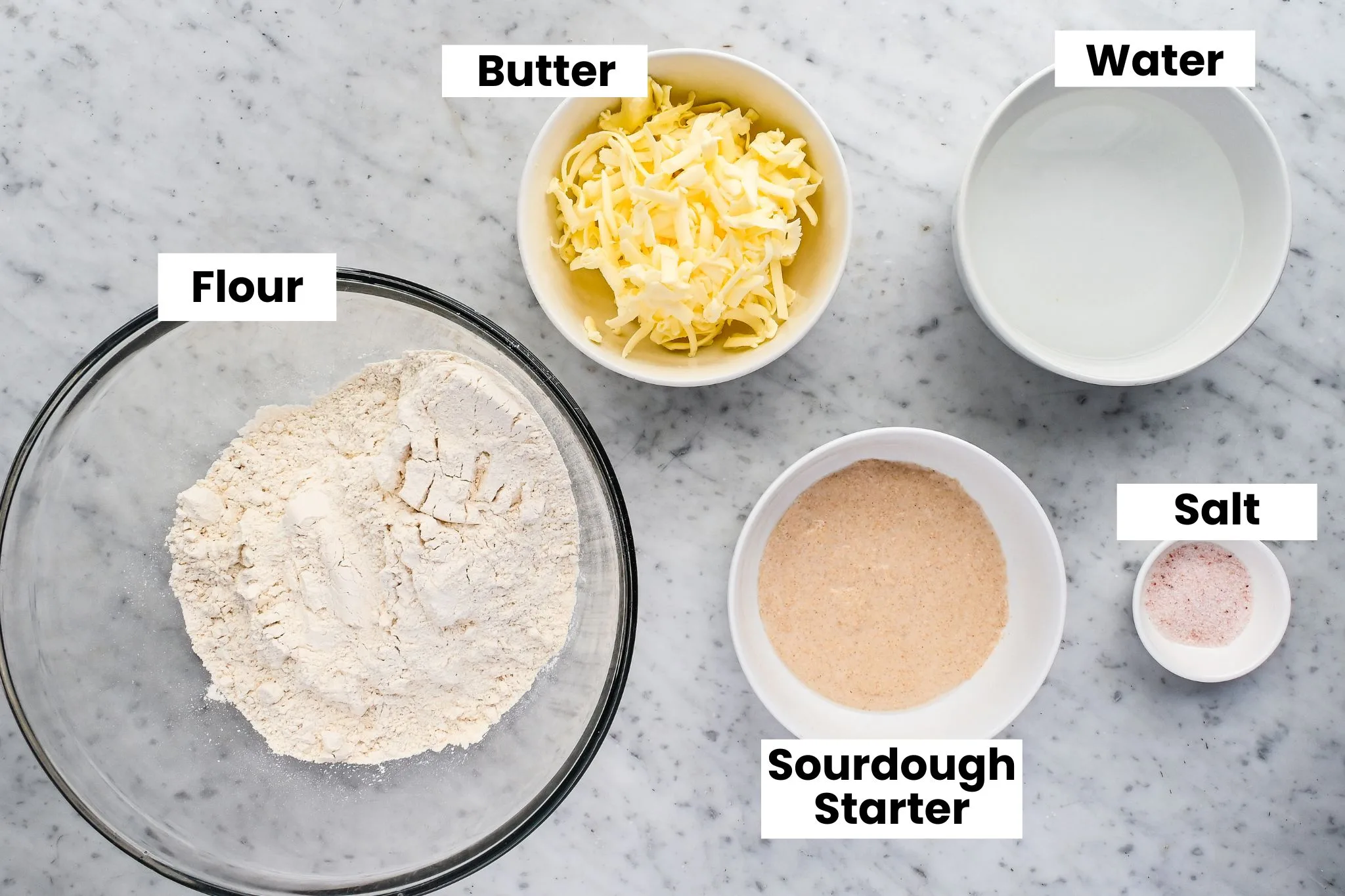 Ingredients for sourdough sandwich bread arranged on a marble surface, including freshly milled flour, butter, water, starter, and salt.
