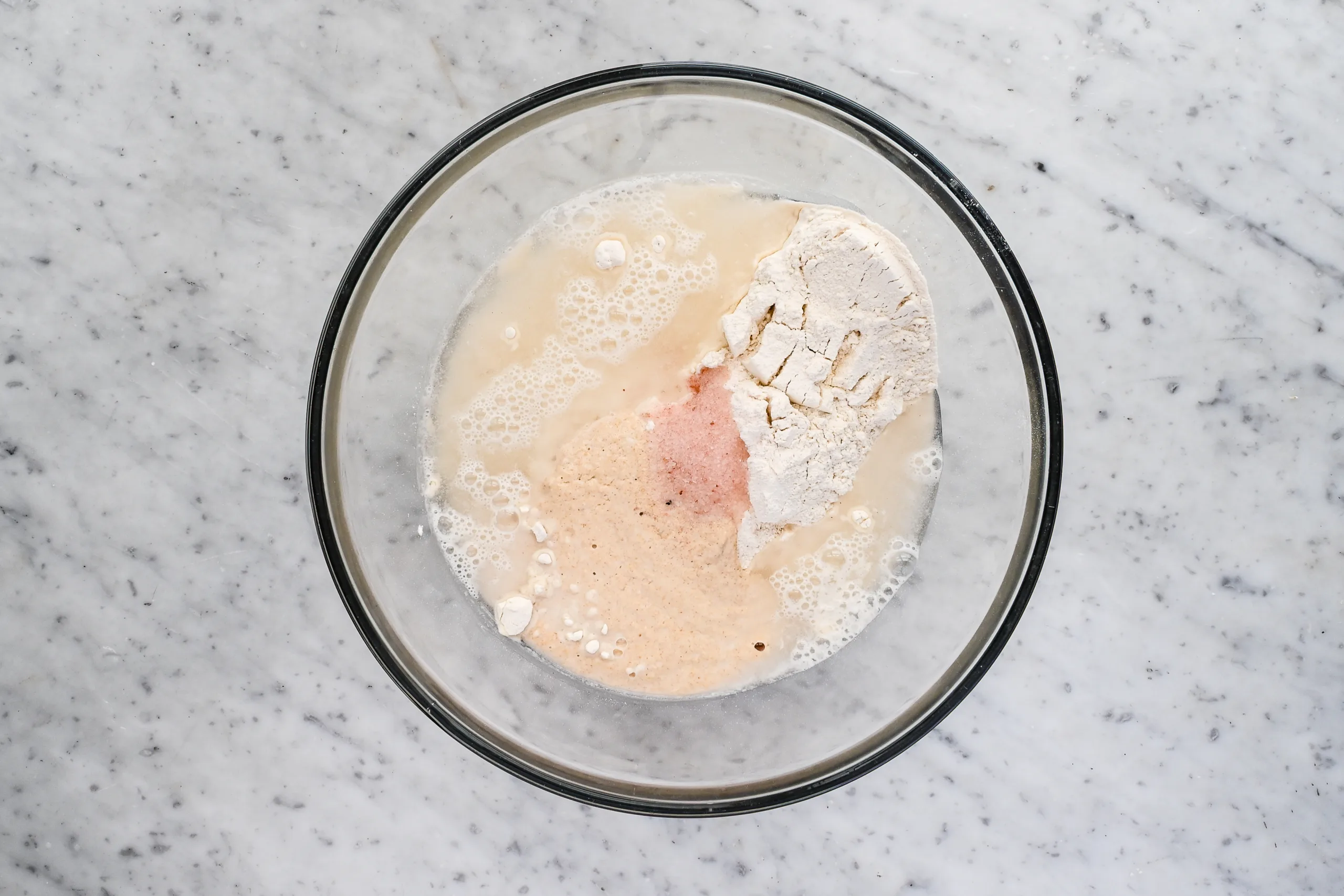 Overhead view of flour, water, salt, and sourdough starter combined in a glass bowl before mixing sandwich bread dough.