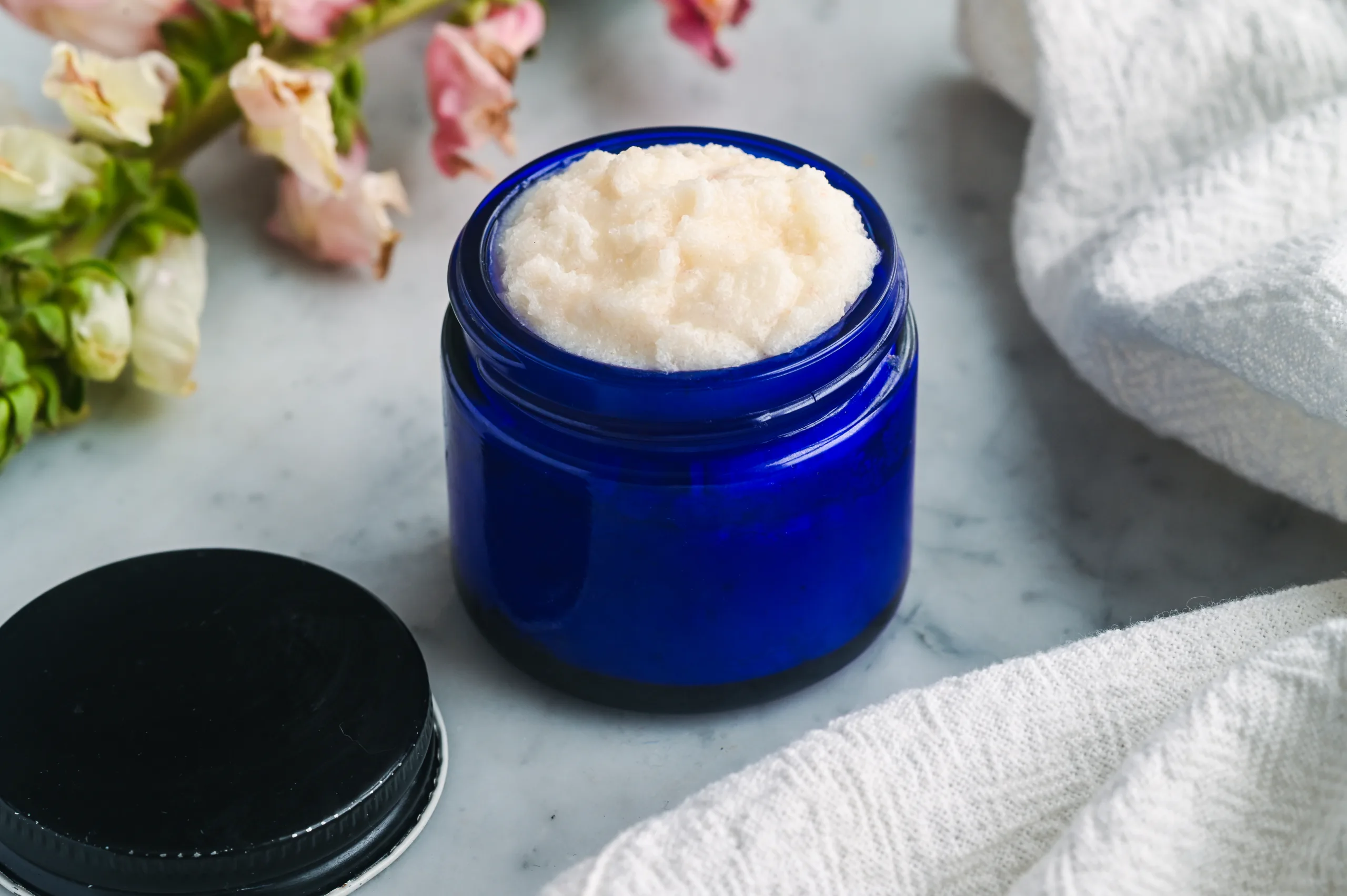 Whipped tallow sugar face scrub with a creamy texture, photographed in a reusable blue glass jar on a marble countertop.