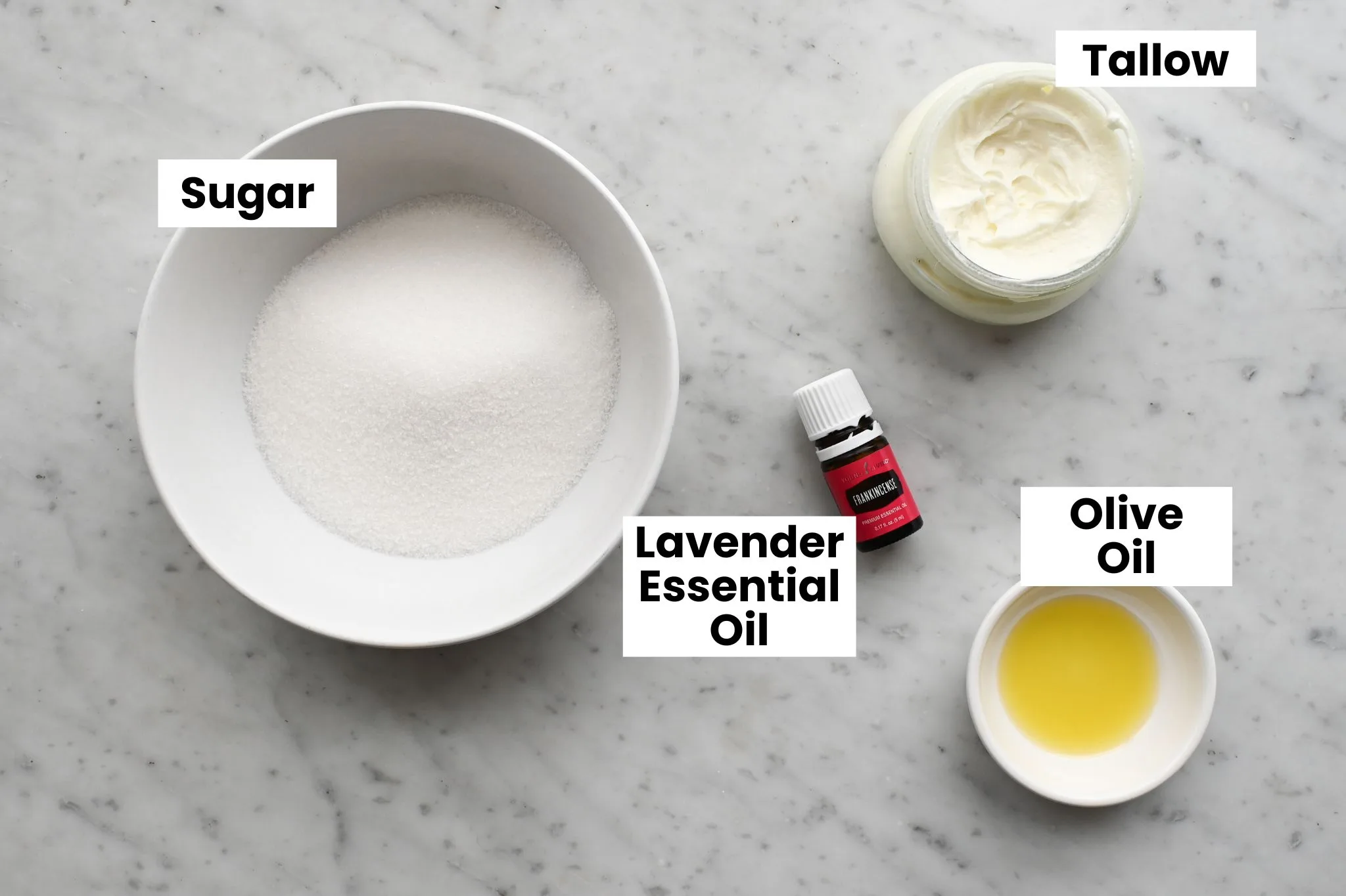 Simple ingredients for a homemade tallow face scrub, including sugar, whipped tallow, olive oil, and lavender essential oil arranged on a marble surface.
