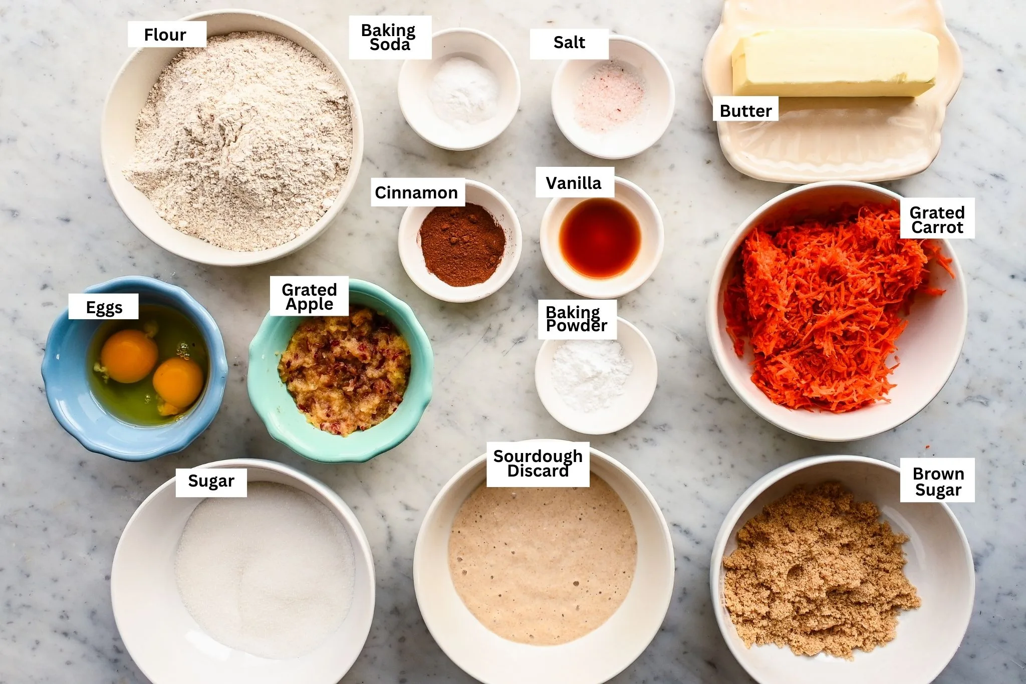 Ingredients for apple carrot sourdough discard breakfast muffins arranged on a marble surface.