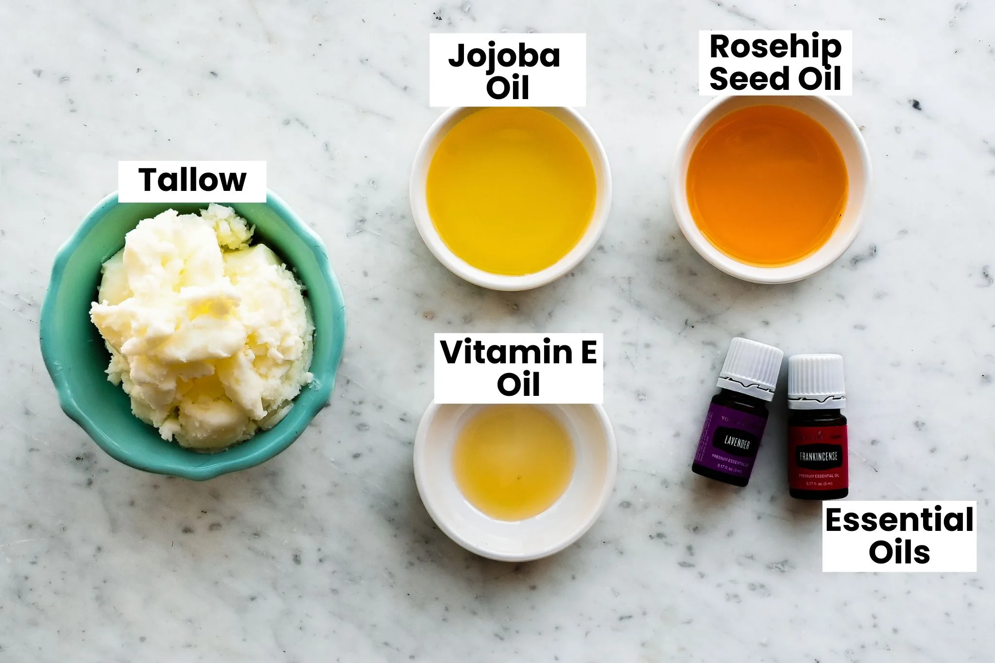 Grass-fed tallow, jojoba oil, rosehip seed oil, vitamin E oil, and essential oils arranged on a marble surface for making a homemade tallow face balm for fine lines and mature skin.