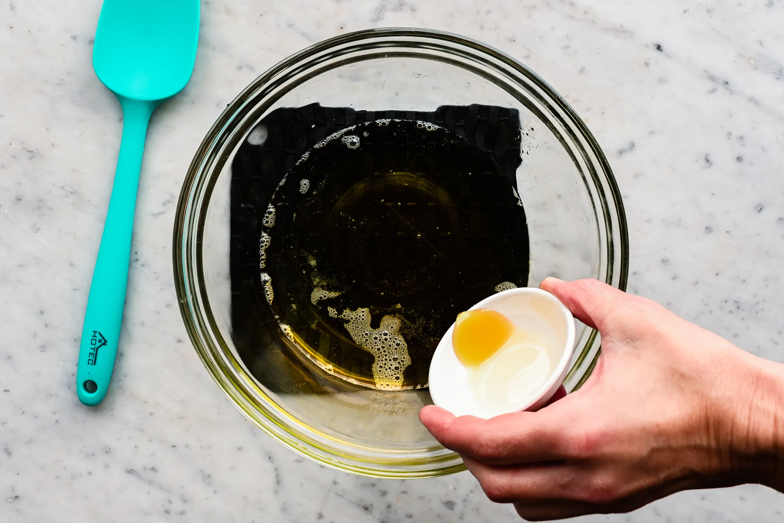 Hand pouring vitamin e oil into melted tallow while mixing ingredients for a diy tallow face cream.