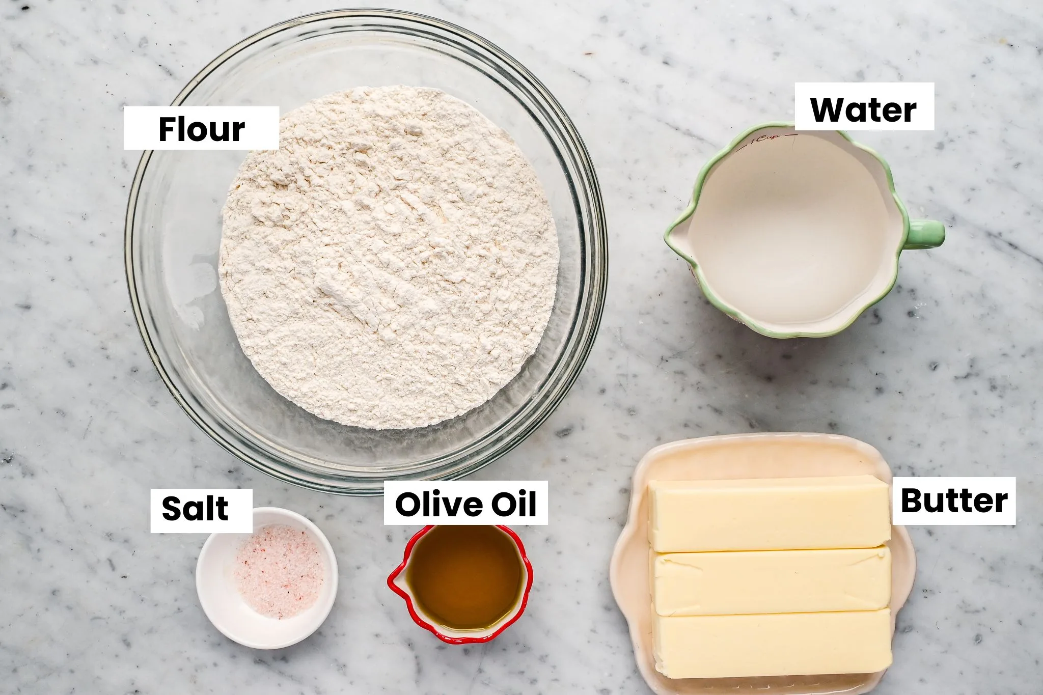 Overhead view of puff pastry ingredients arranged on a marble surface, including flour in a glass bowl, water in a measuring cup, butter, olive oil, and salt labeled for an old-fashioned homemade puff pastry recipe.
