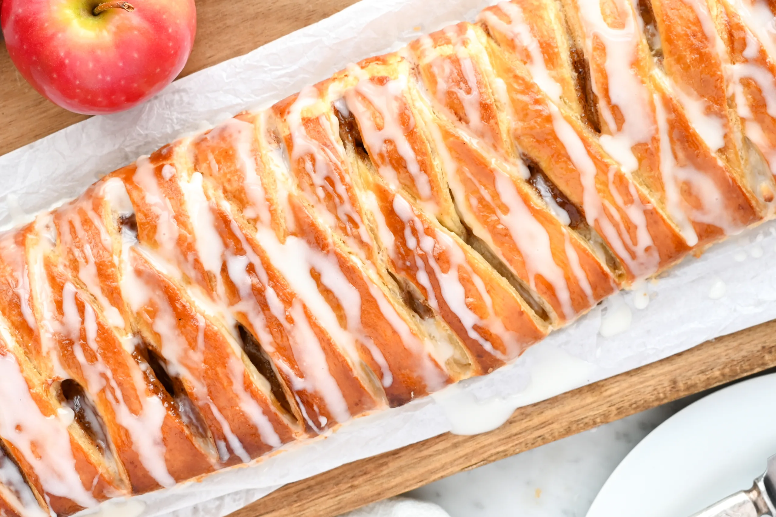 Braided apple cream cheese puff pastry Danish drizzled with sweet glaze, served on parchment paper with fresh red apples nearby.