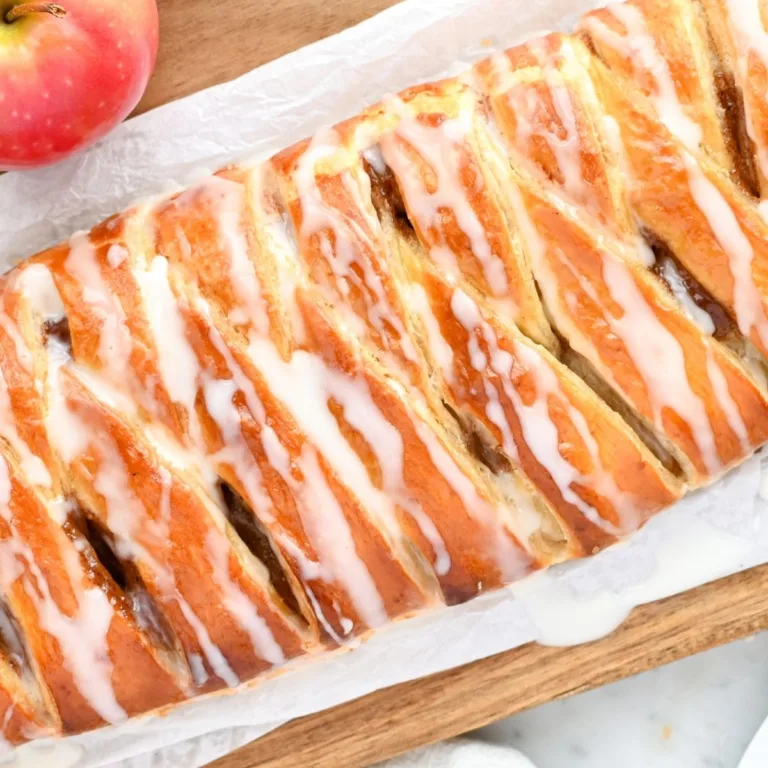 Golden braided apple cream cheese puff pastry Danish topped with a sweet vanilla glaze, showing flaky layers and cinnamon apple filling peeking through the folds, served on parchment over a wooden board with fresh apples nearby.