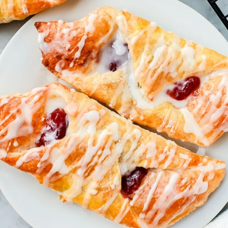 Golden old-fashioned strawberry Danish pastries made with homemade puff pastry, folded into triangles and filled with bright red strawberry jam, then drizzled with a simple vanilla glaze and served on a white plate.