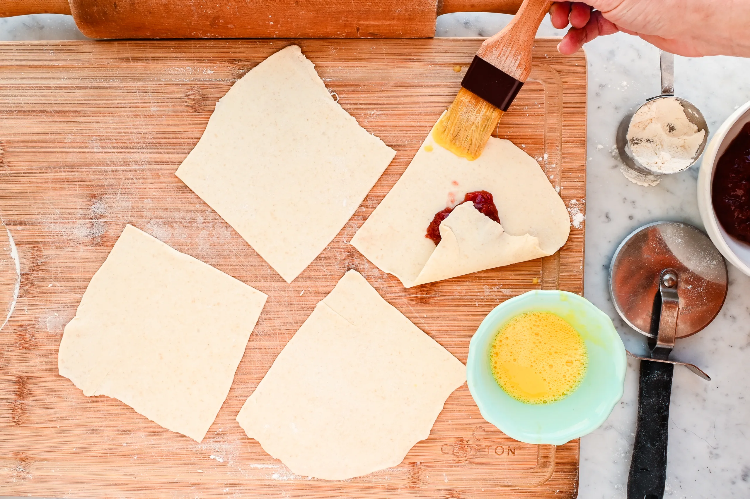Brushing egg wash onto puff pastry square filled with strawberry jam before folding into Danish shape.