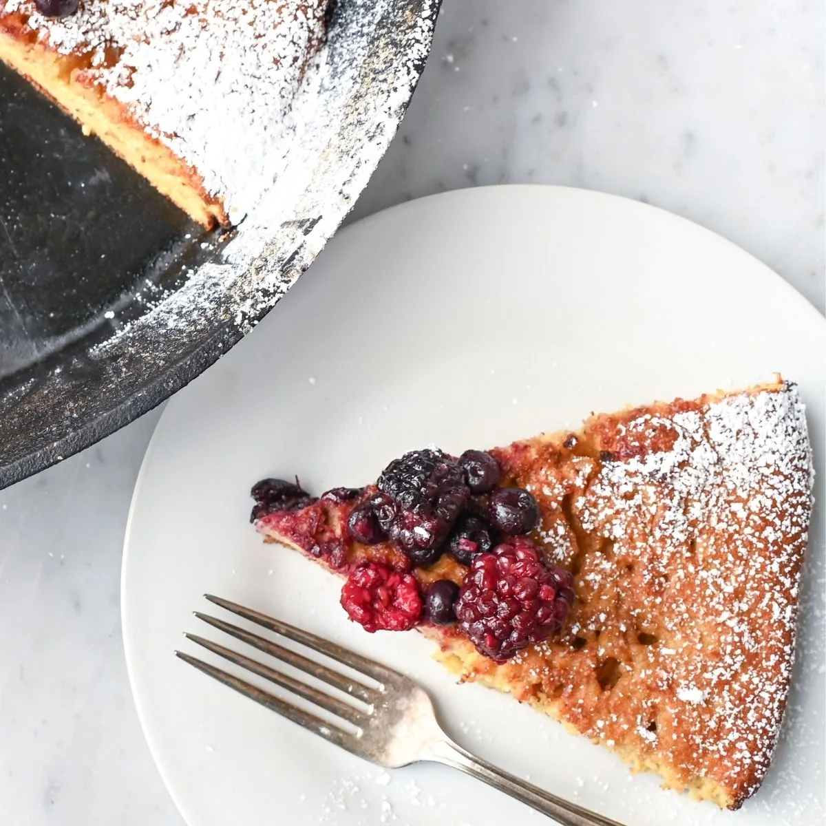 Slice of sourdough German pancake with fresh berries and powdered sugar served on a white plate with a fork.