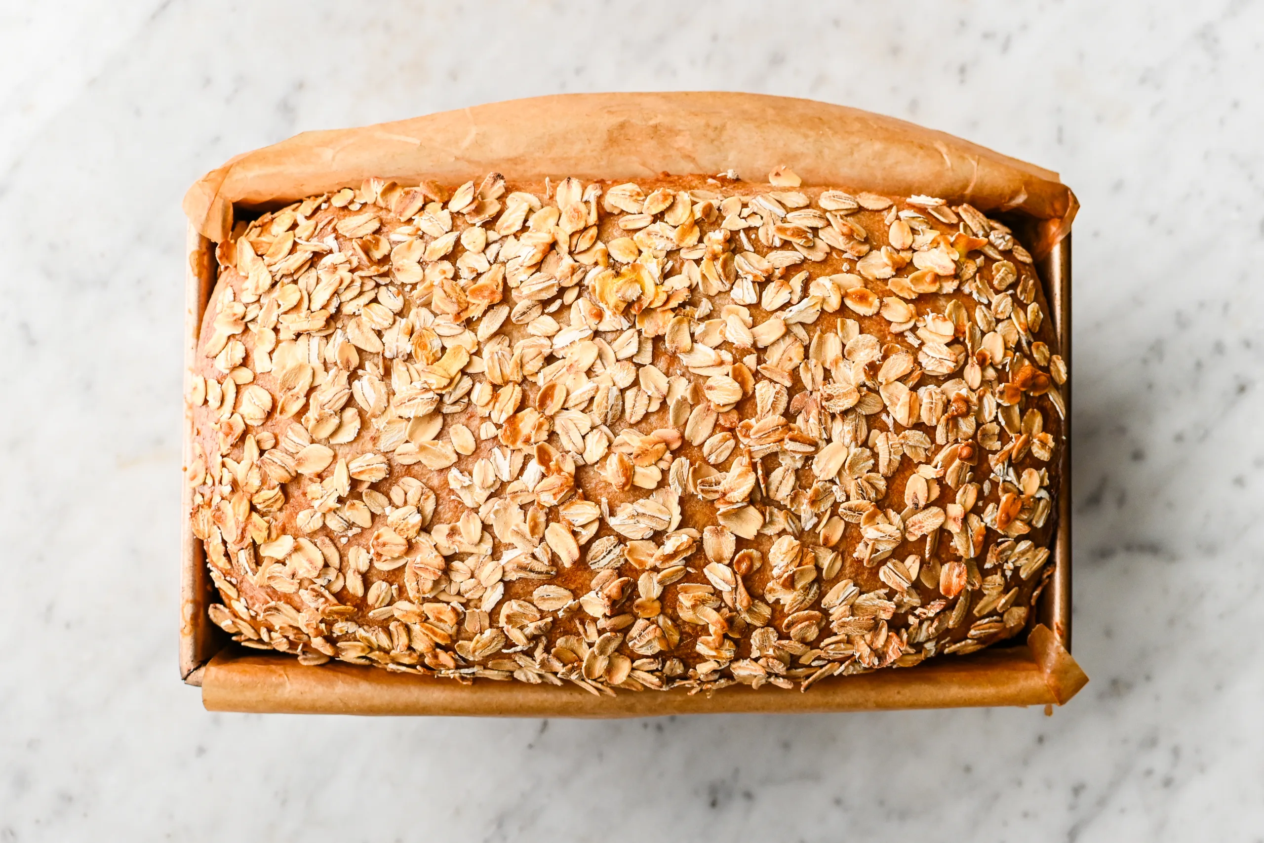 Freshly baked sourdough oat sandwich loaf topped with toasted rolled oats in a parchment-lined loaf pan, viewed from above.