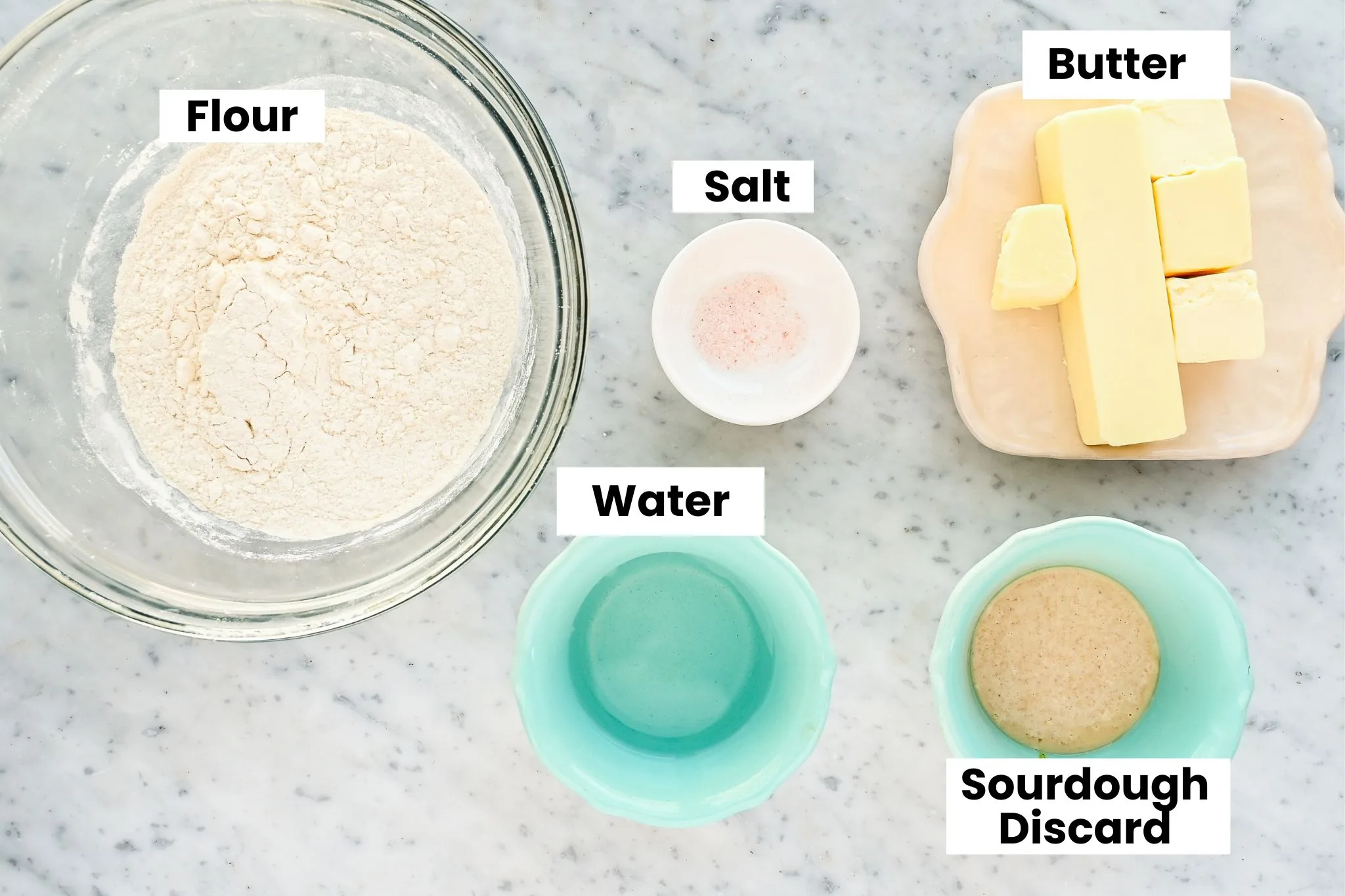 Ingredients for sourdough puff pastry including flour, butter, salt, water, and sourdough starter arranged on a marble countertop.