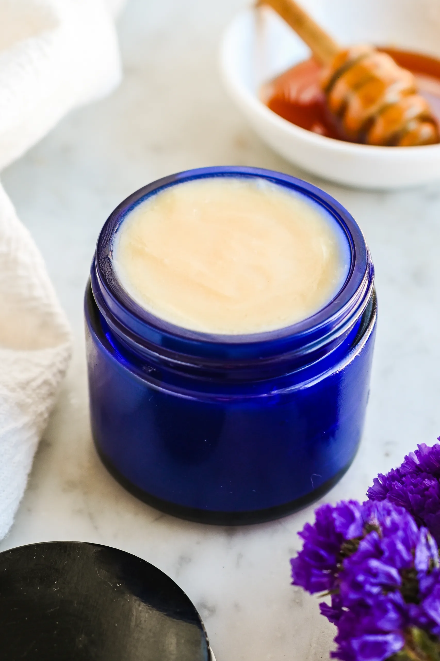 Old-fashioned tallow and honey balm in a blue jar styled with honey dipper and soft linens on a marble surface.