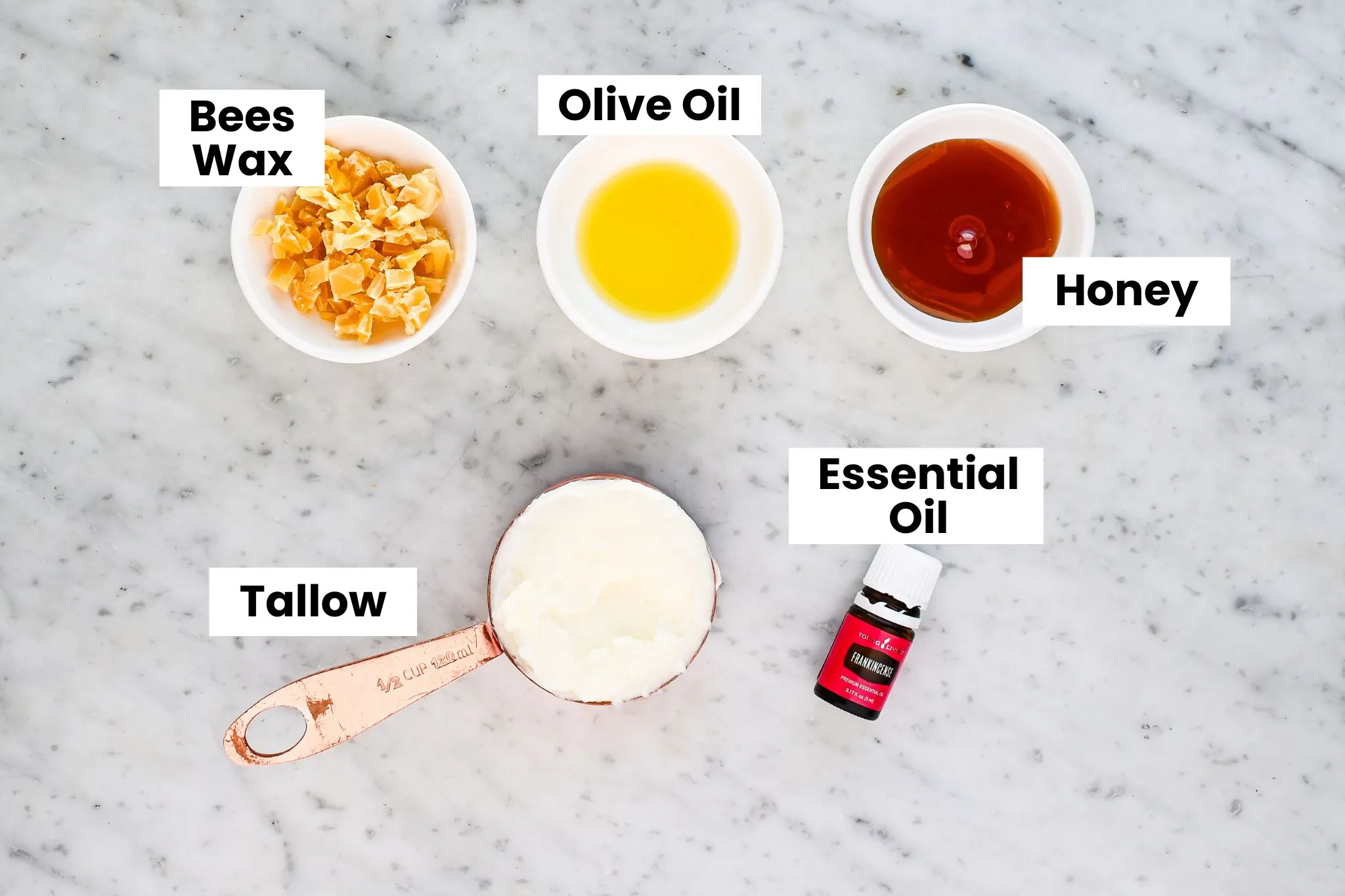 Ingredients for old-fashioned tallow honey balm including beeswax, olive oil, honey, grass fed beef tallow, and essential oil arranged on a marble surface.