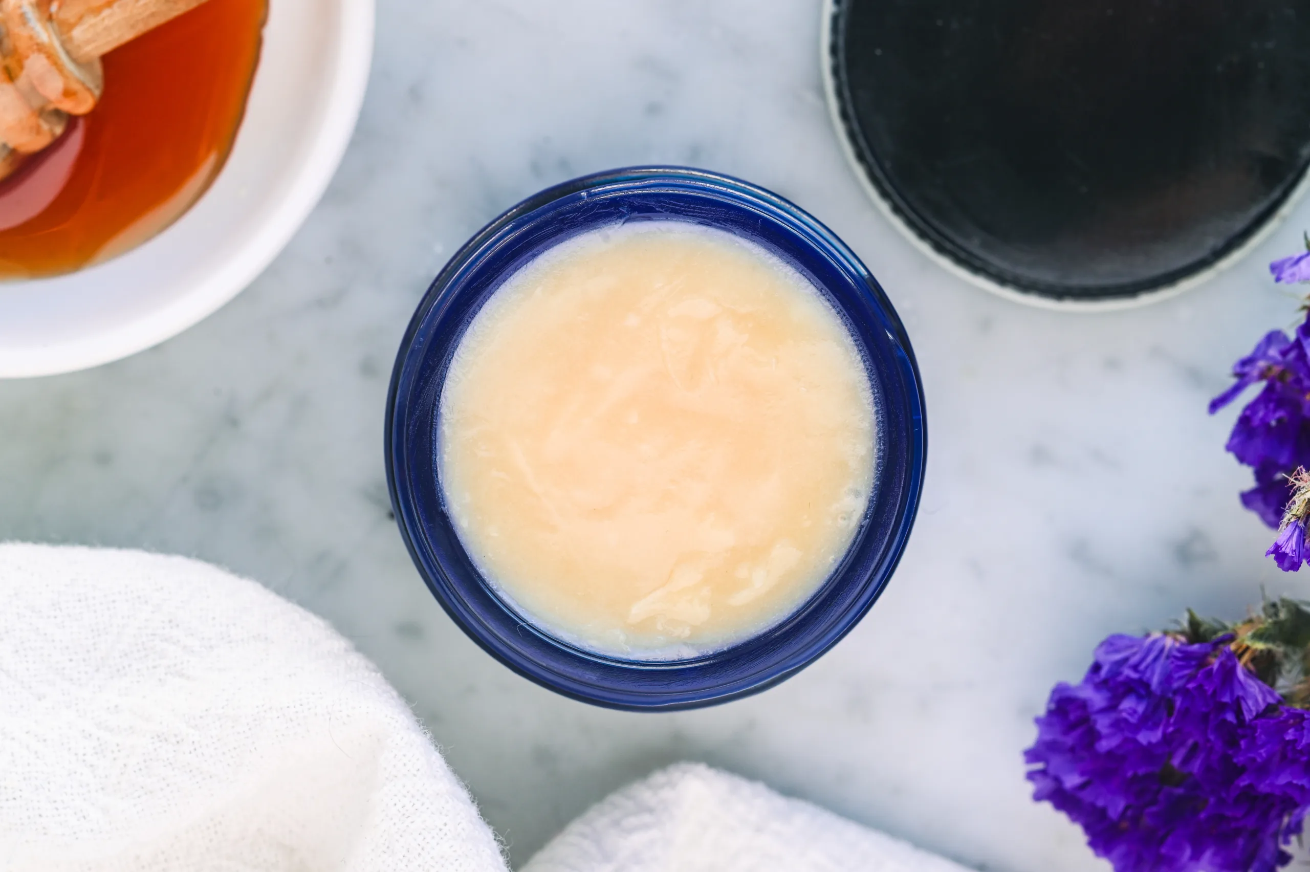 Finished tallow and honey balm set in a blue glass jar with a smooth creamy texture, surrounded by honey and flowers.