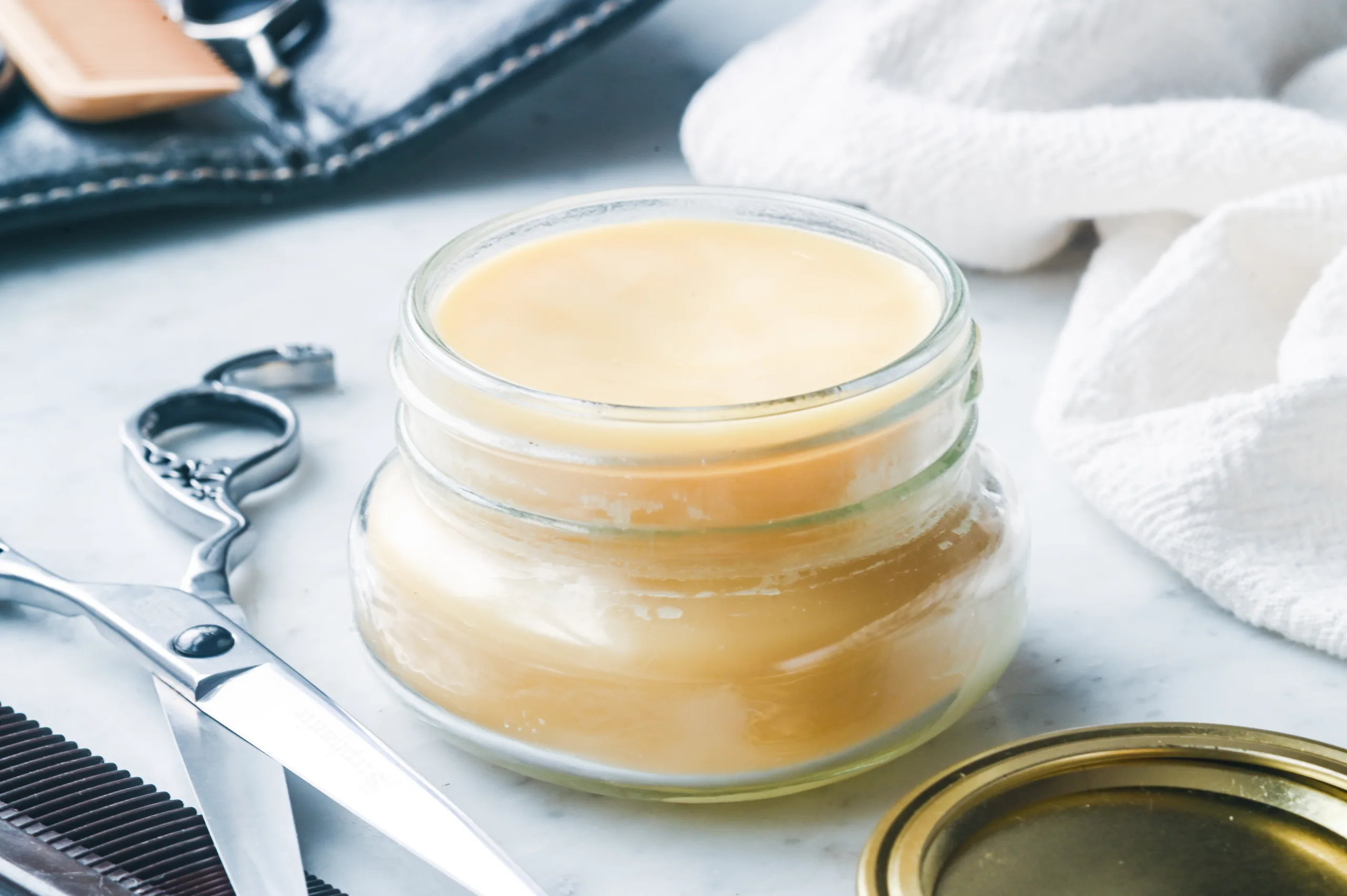 Finished homemade natural pomade in a small glass jar on a countertop with grooming tools.