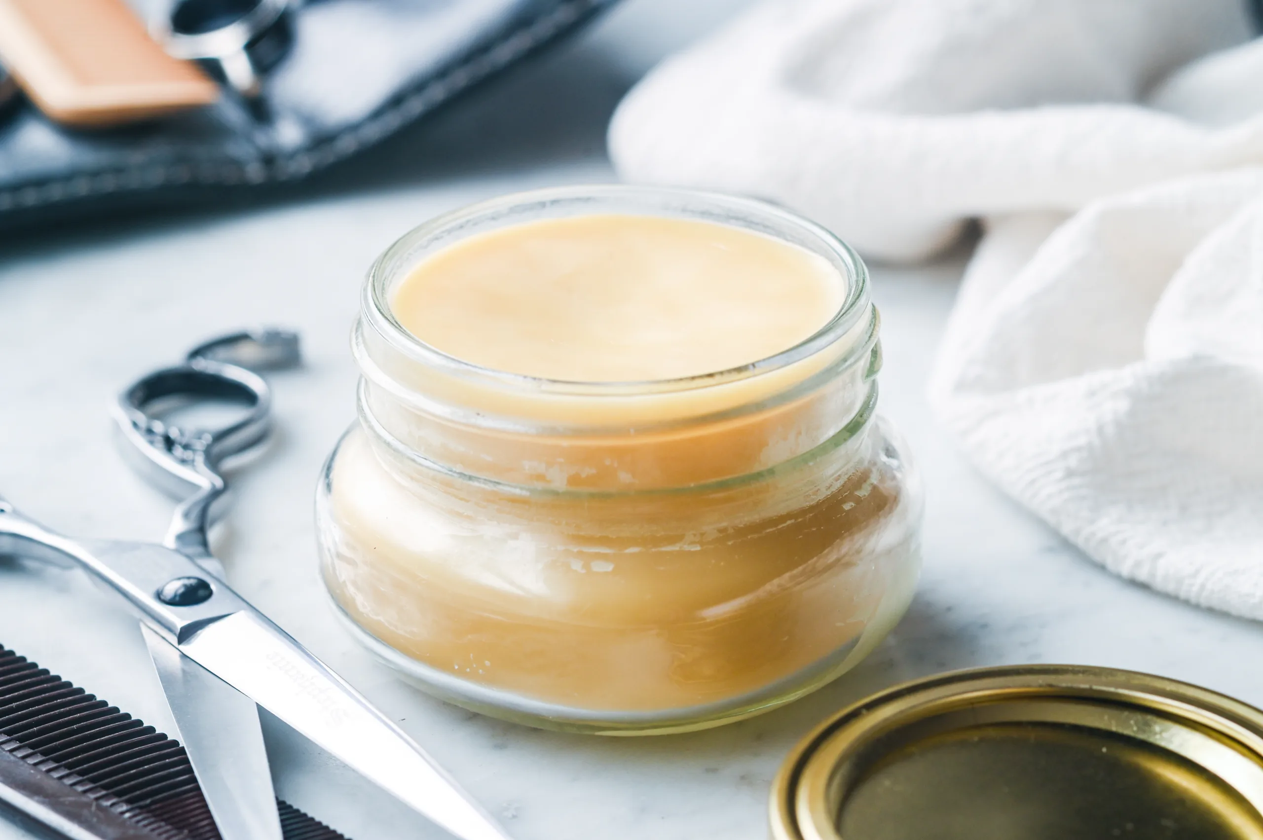 Homemade beeswax and tallow pomade in a glass jar beside scissors and a comb.