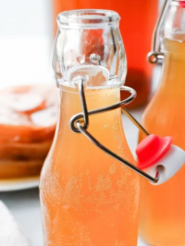 Homemade kombucha in clear swing-top bottles with light fizz bubbles visible, styled on a bright surface with a SCOBY blurred in the background.