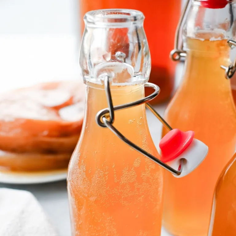 Homemade kombucha in clear swing-top bottles with light fizz bubbles visible, styled on a bright surface with a SCOBY blurred in the background.