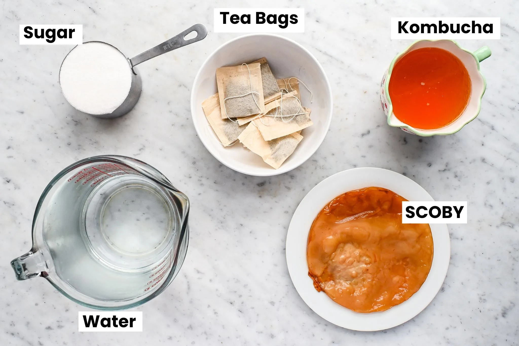 Ingredients for homemade kombucha on a marble countertop, including sugar, tea bags, water, starter tea, and a kombucha scoby.