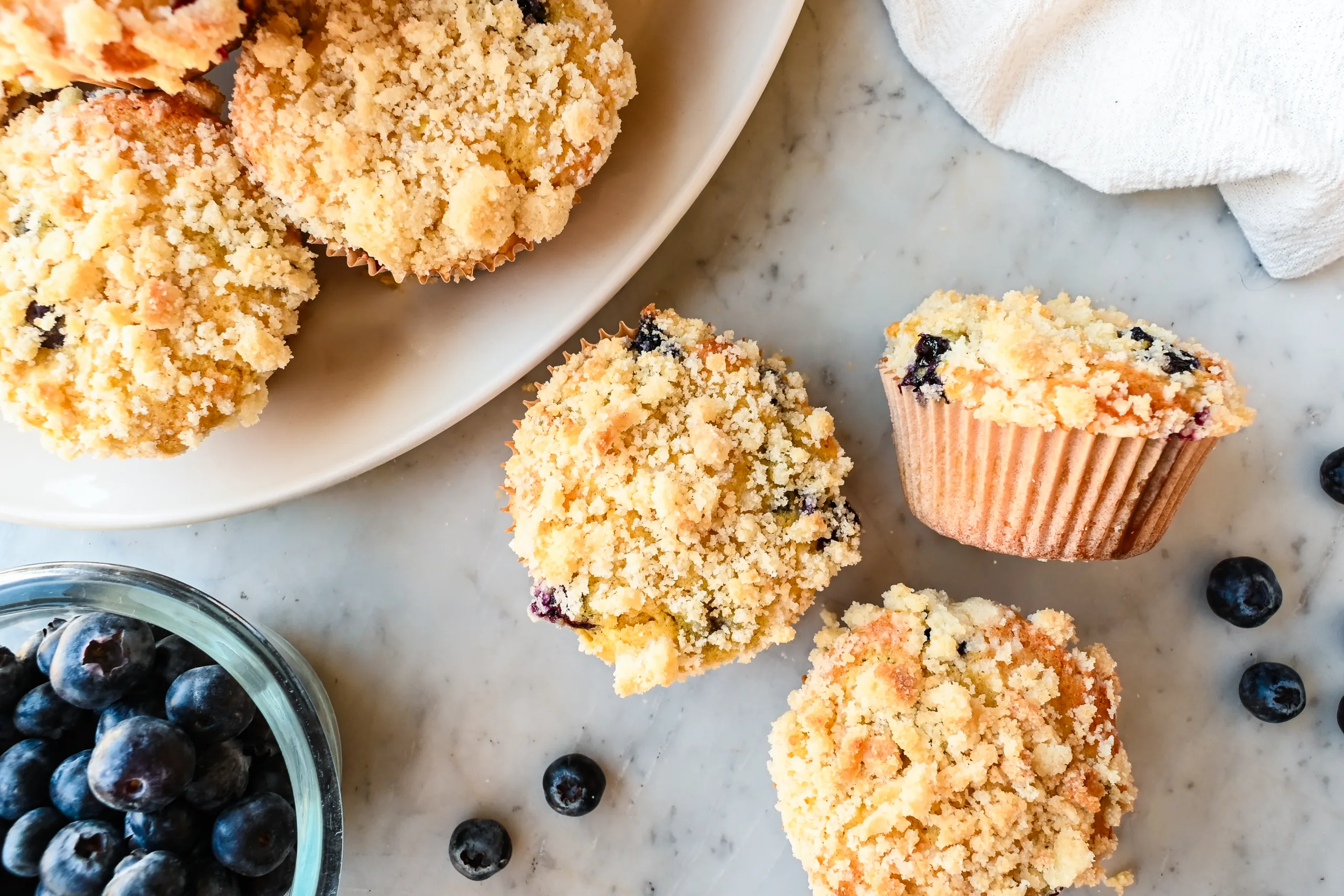 Three sourdough discard blueberry muffins with crumb topping on a marble countertop beside a bowl of fresh blueberries and a white plate.