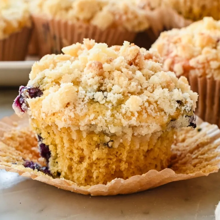 Close-up of a sourdough discard blueberry muffin in its paper liner, topped with buttery crumb topping and filled with juicy blueberries.
