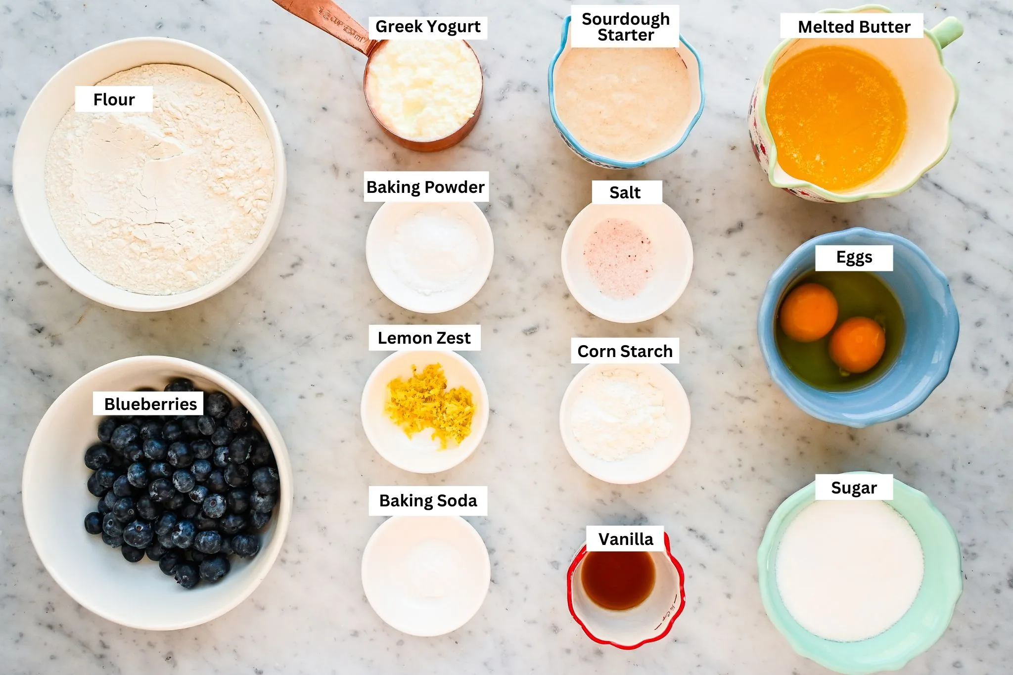 Ingredients for sourdough discard blueberry muffins with crumb topping arranged on a marble countertop, including flour, blueberries, sourdough discard, melted butter, sugar, eggs, vanilla, lemon zest, salt, baking powder, and cornstarch.
