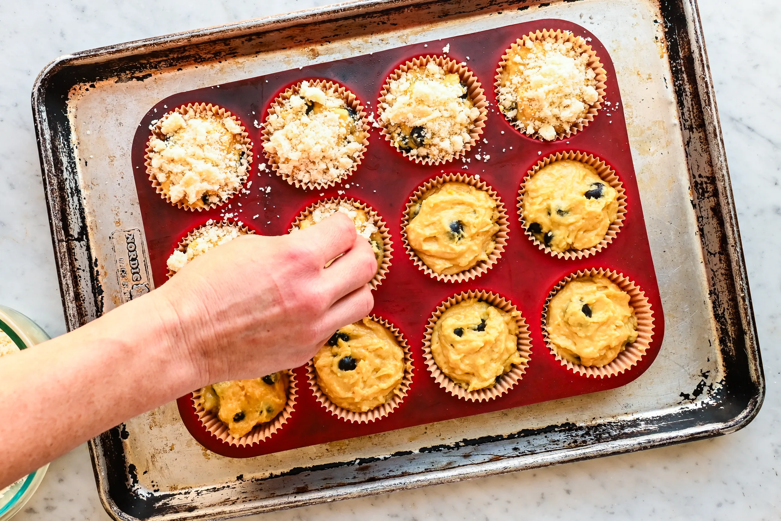 Hand sprinkling crumb topping over sourdough discard blueberry muffin batter in paper liners set in a red silicone muffin pan on a baking sheet.