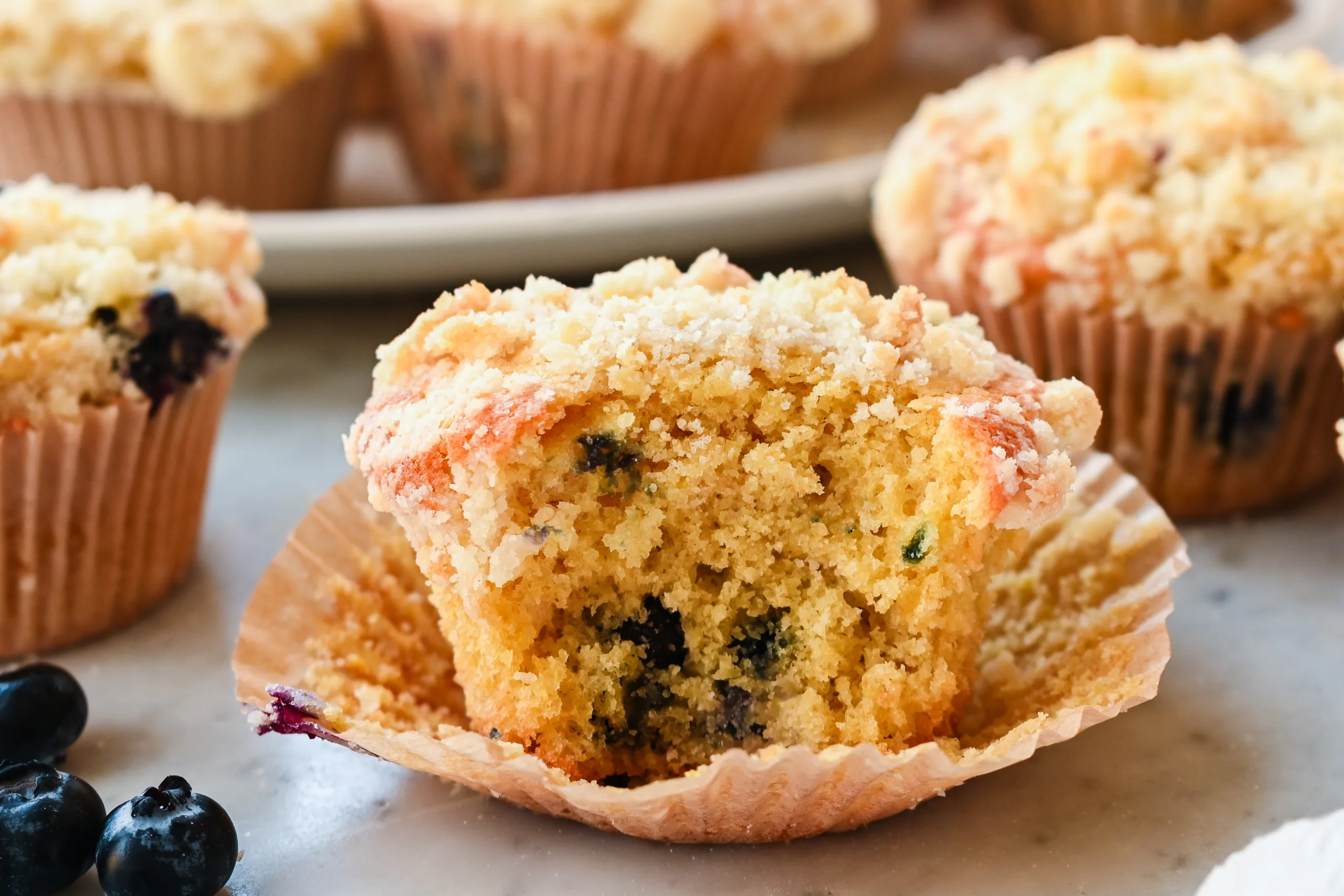 Sourdough discard blueberry muffin with crumb topping opened in its paper liner to show the soft, tender interior filled with blueberries.