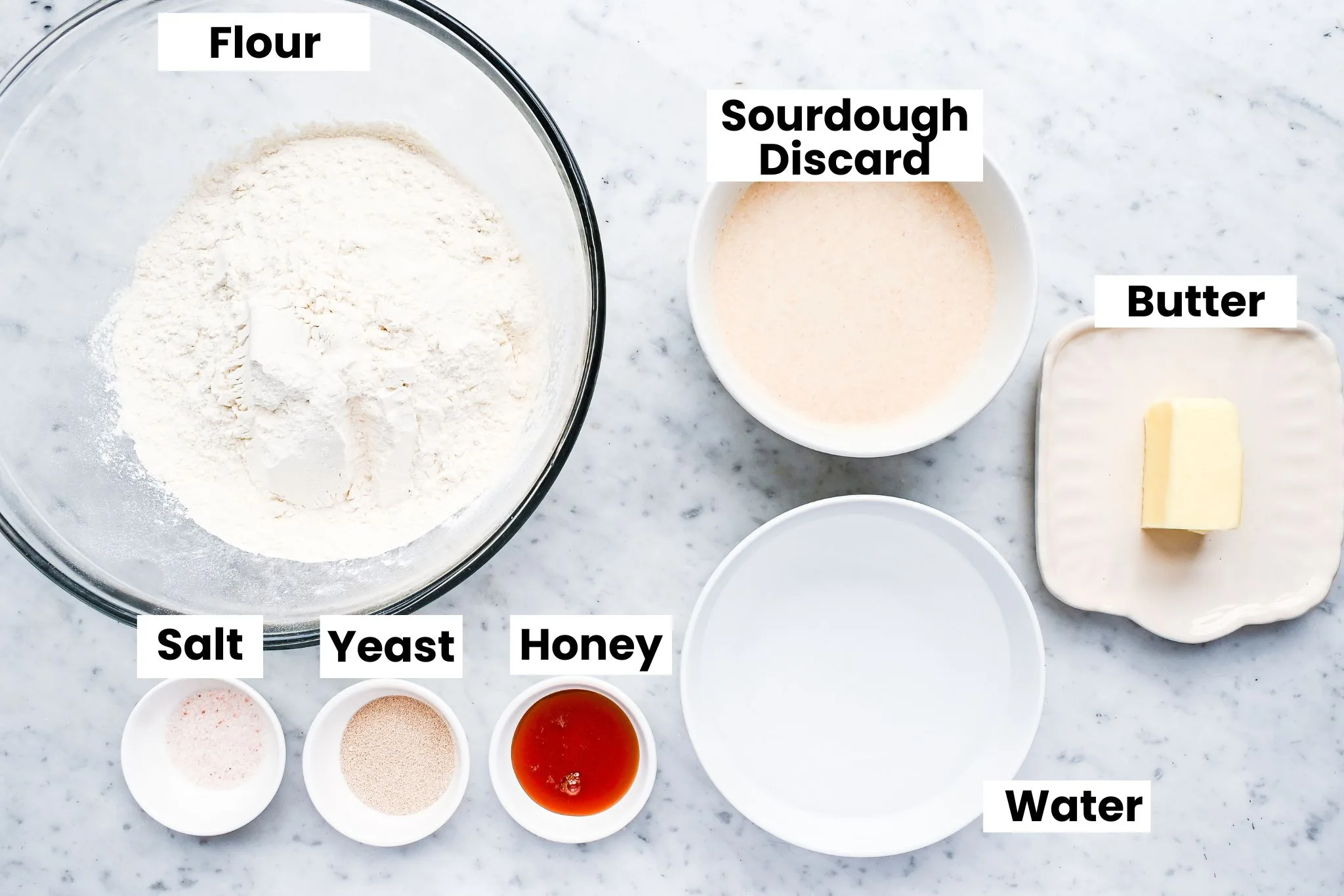 Ingredients for sourdough discard sandwich bread arranged on a marble countertop including flour, yeast, sugar, butter, salt, and water.
