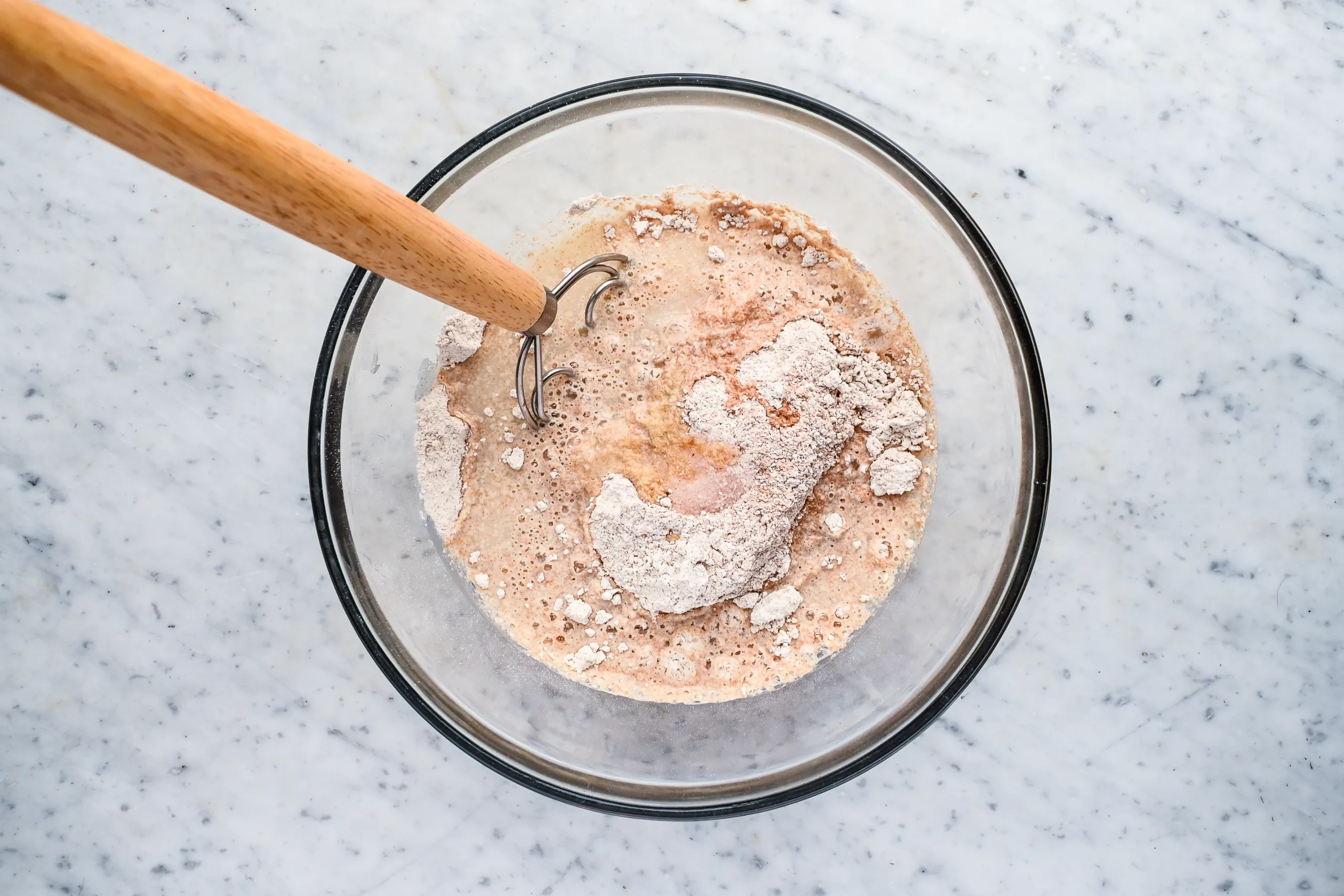 Sourdough starter, honey, and water being added to fresh milled spelt flour in a glass bowl.