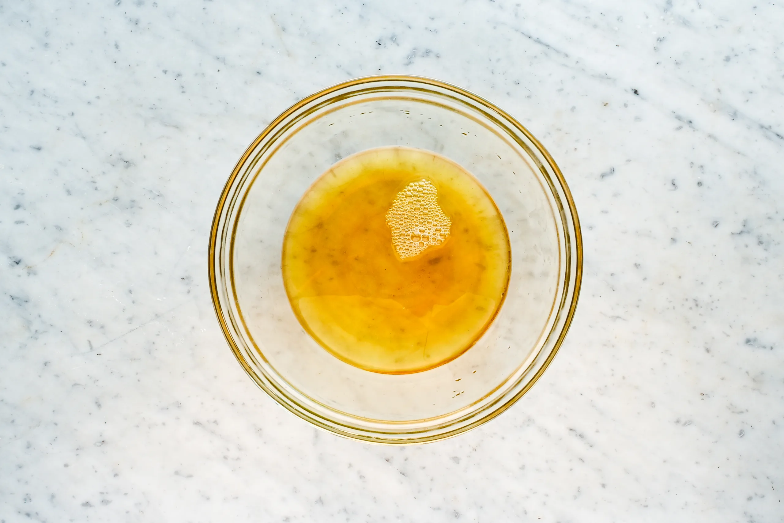 Melted tallow, shea butter, and cocoa butter in a glass bowl on a marble countertop before whipping.