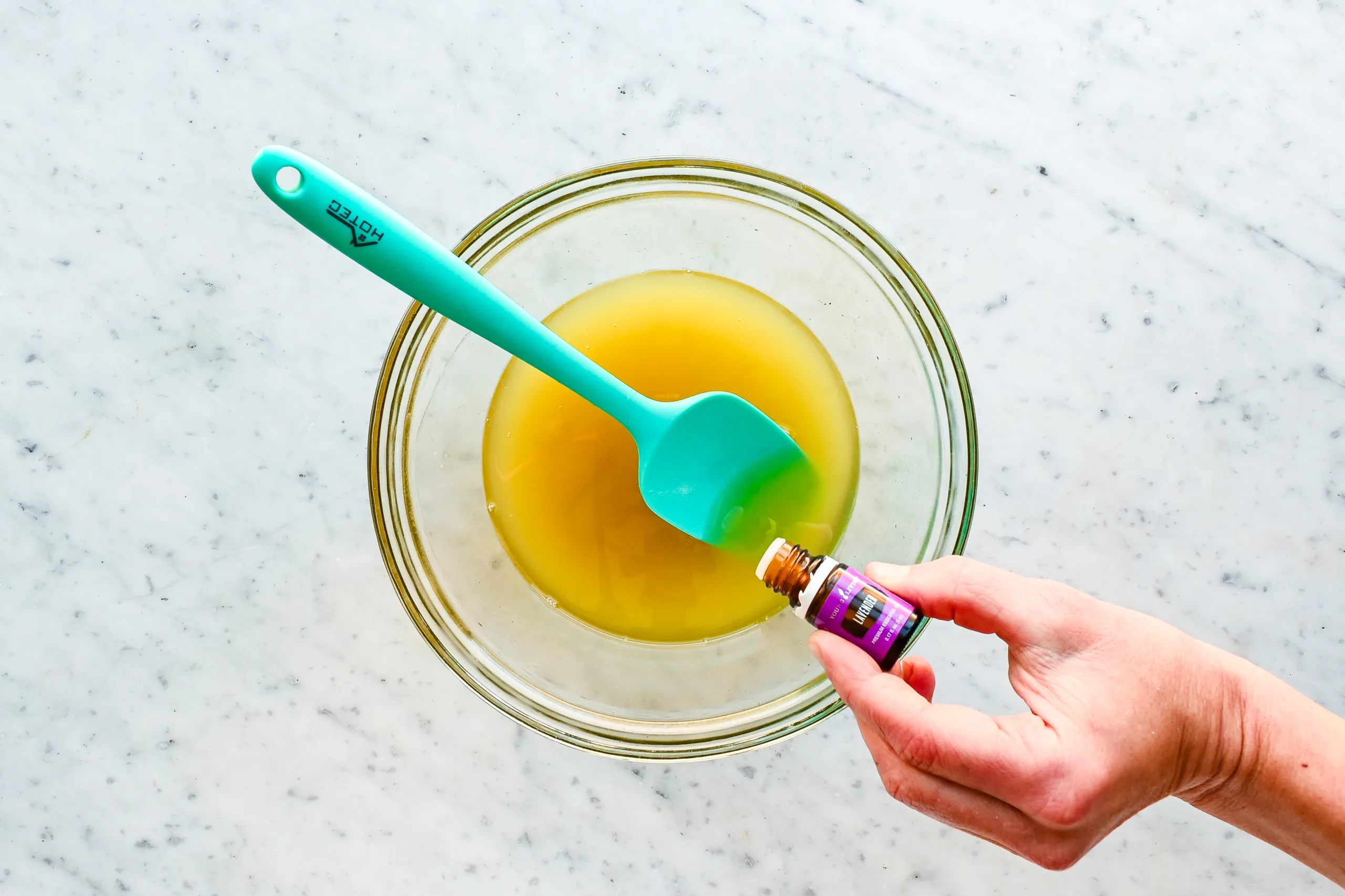 Hand adding lavender essential oil to melted tallow body butter mixture in a glass bowl with a silicone spatula.