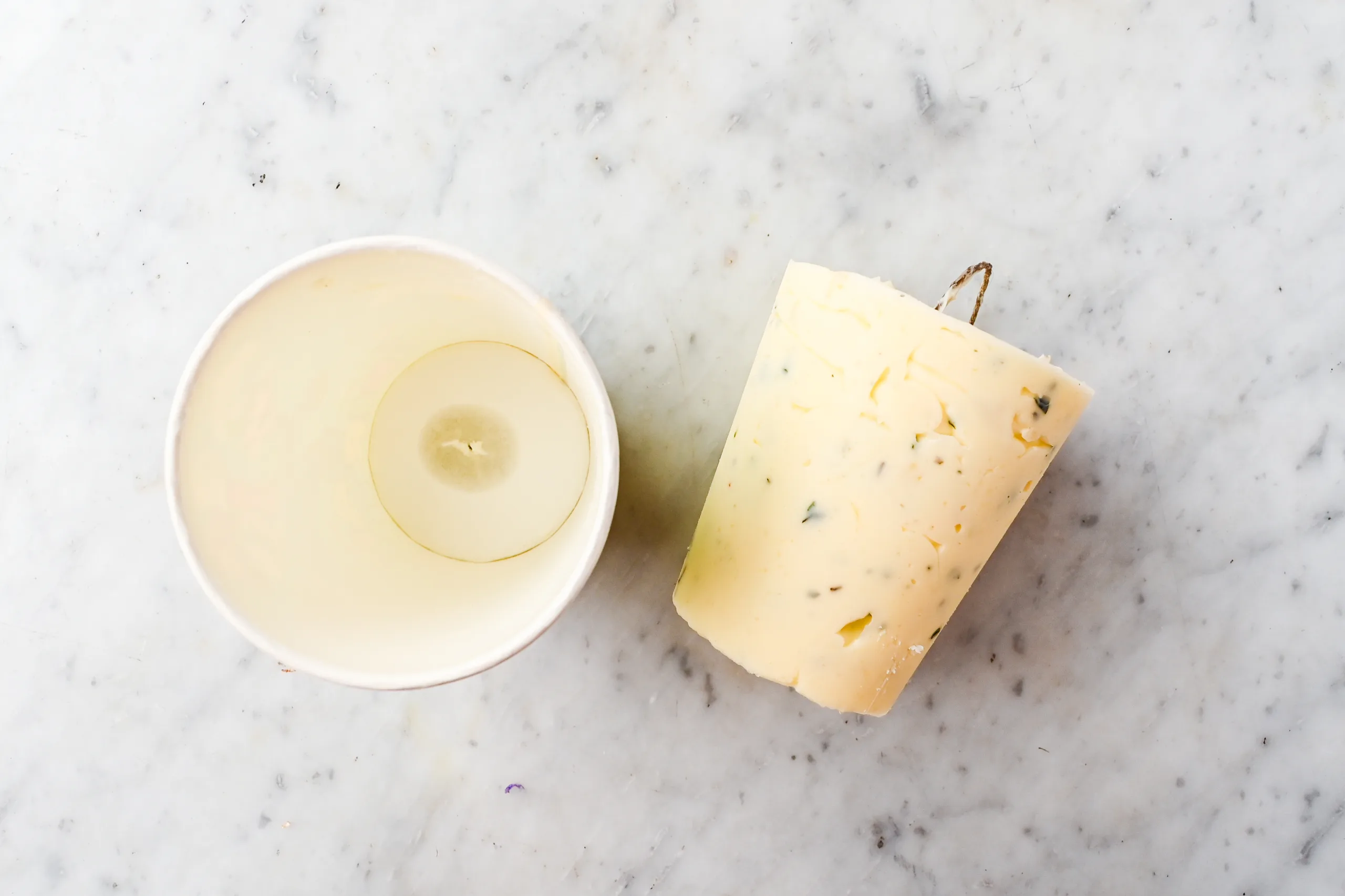 Paper cup removed from an herb butter candle, showing the smooth sides of the finished candle and the inside of the cup beside it on a marble surface