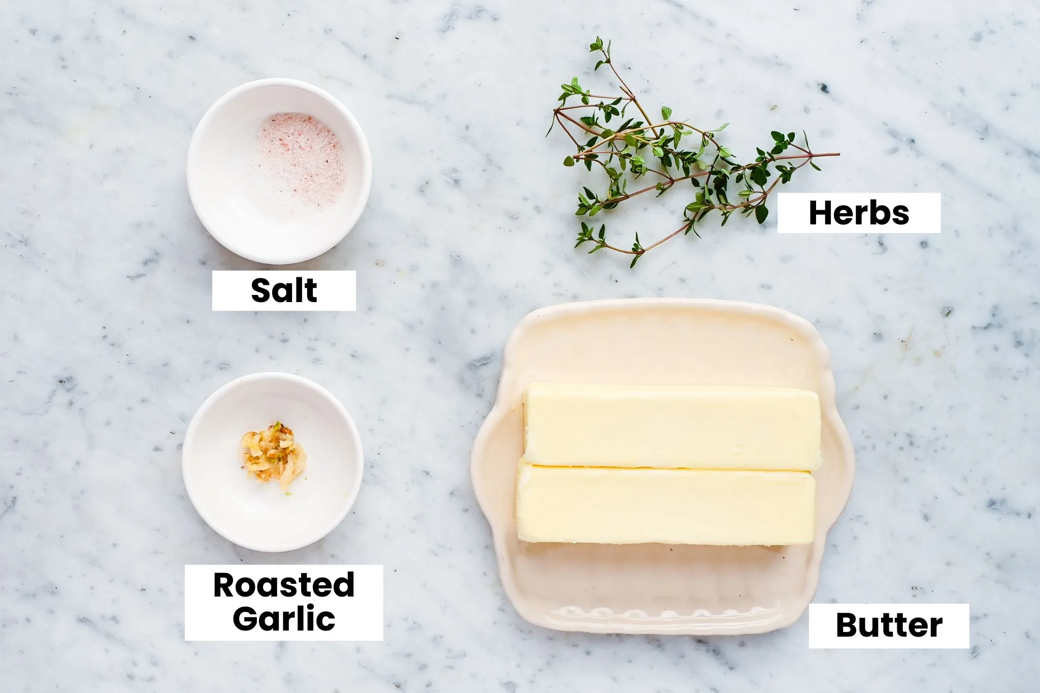 Ingredients for a garlic butter candle recipe labeled on a marble surface, including butter, roasted garlic, salt, and fresh herbs