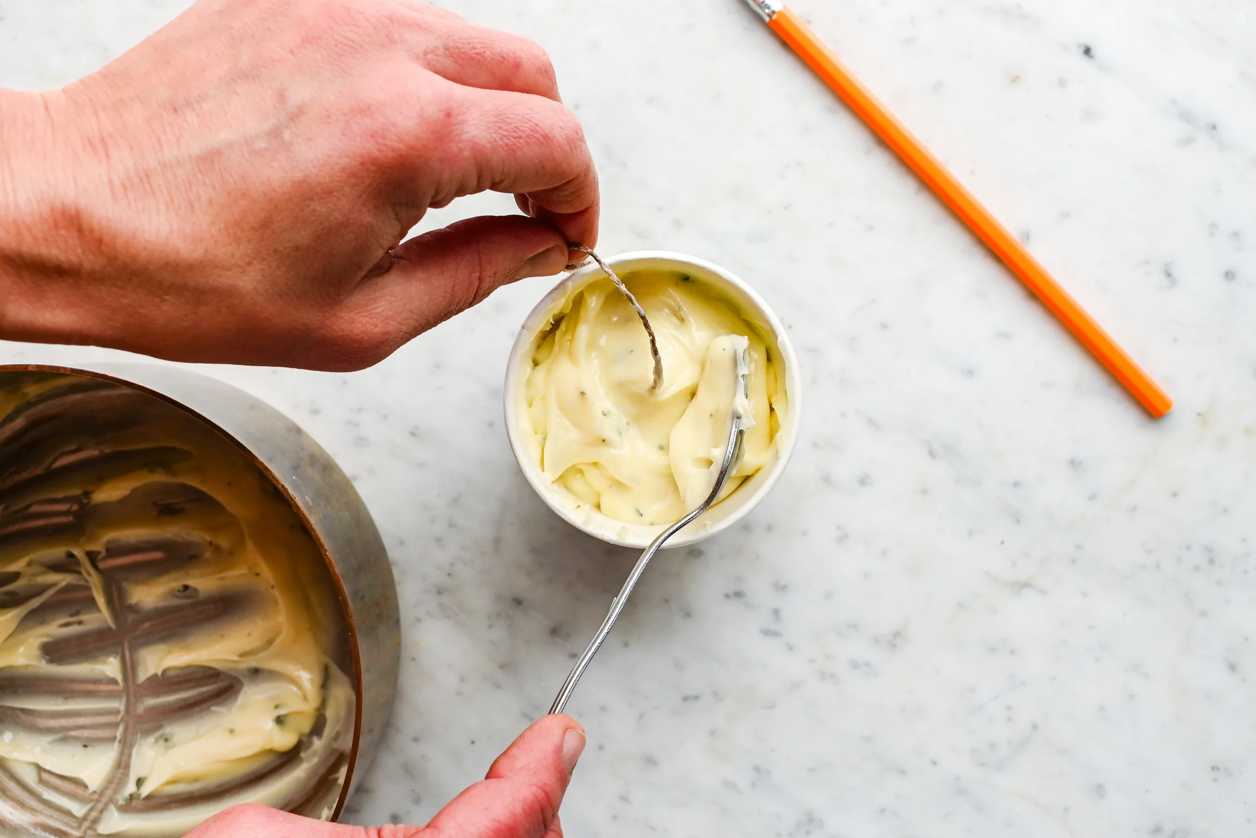 Garlic herb butter mixture being spooned into a paper cup around a centered wick to form a butter candle.