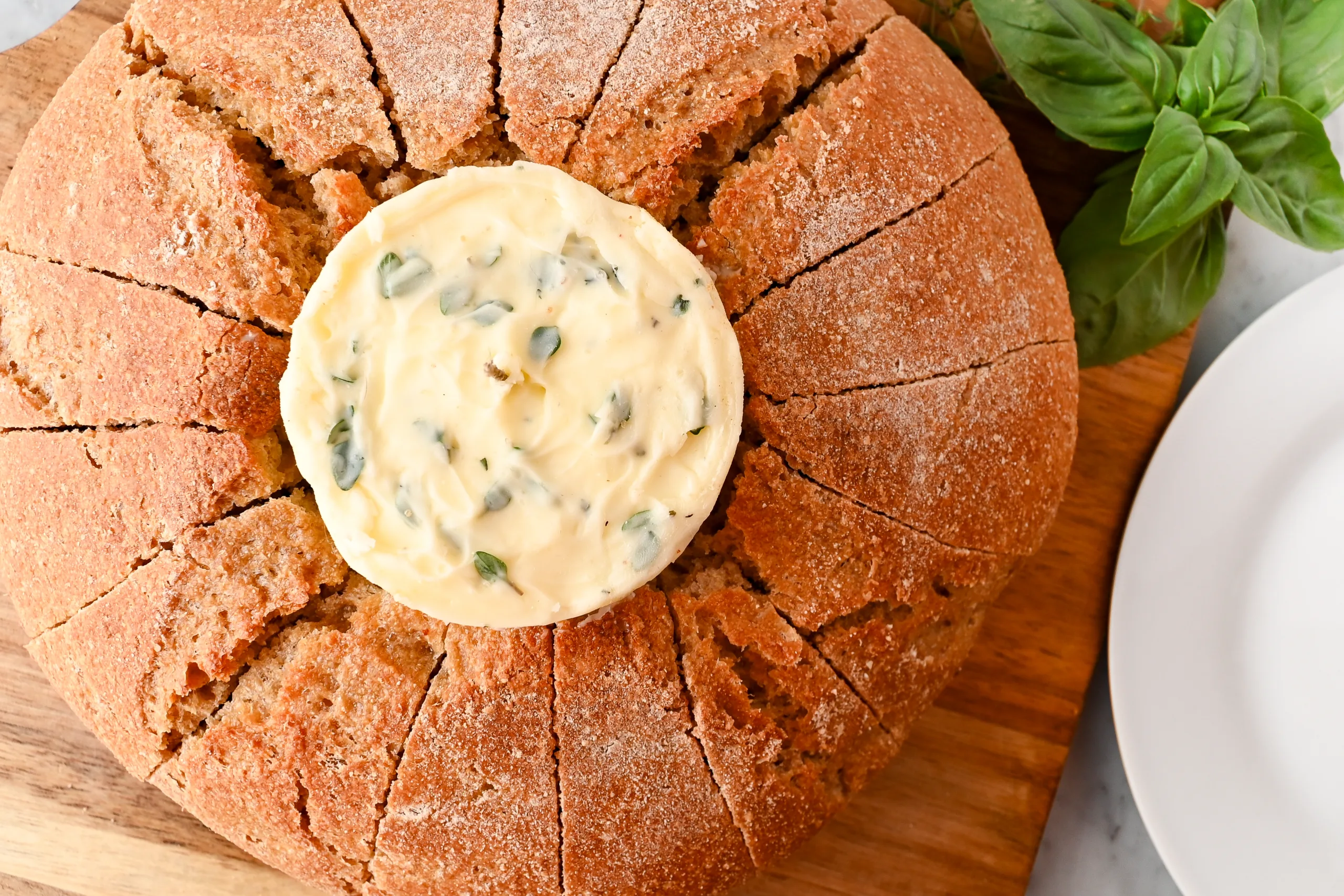 Round loaf of bread cut into slices around an unlit garlic butter candle, styled on a wooden board with fresh basil.