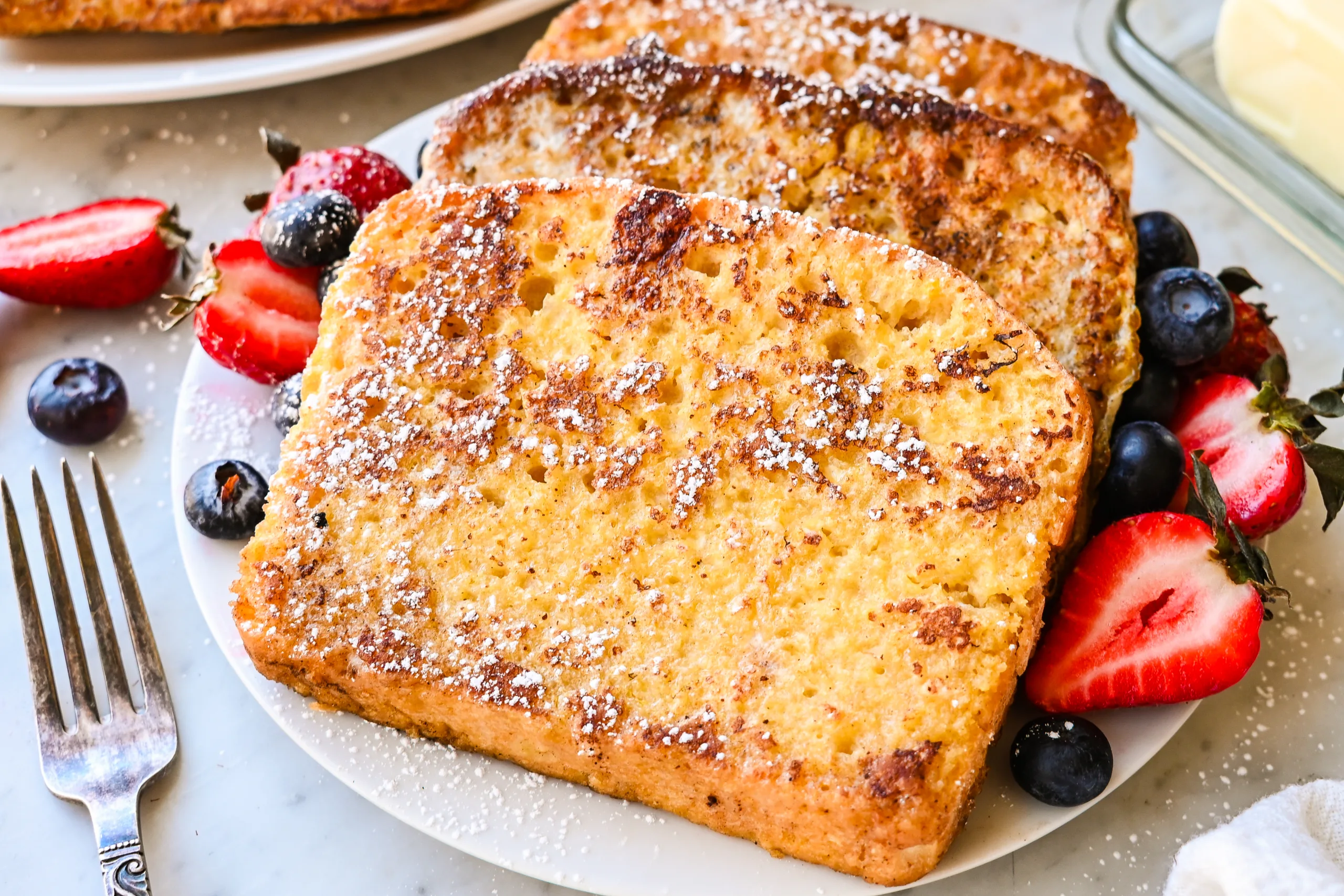Close-up plate of sourdough French toast with powdered sugar, strawberries, and blueberries.